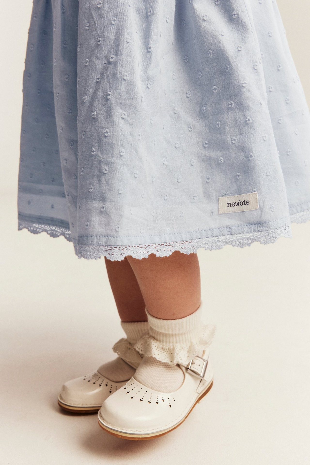Nahaufnahme eines Kindes in hellblauem gepunktetem Kleid mit Spitzenbesatz, weißen Rüschensocken und weißen Schnallenschuhen.