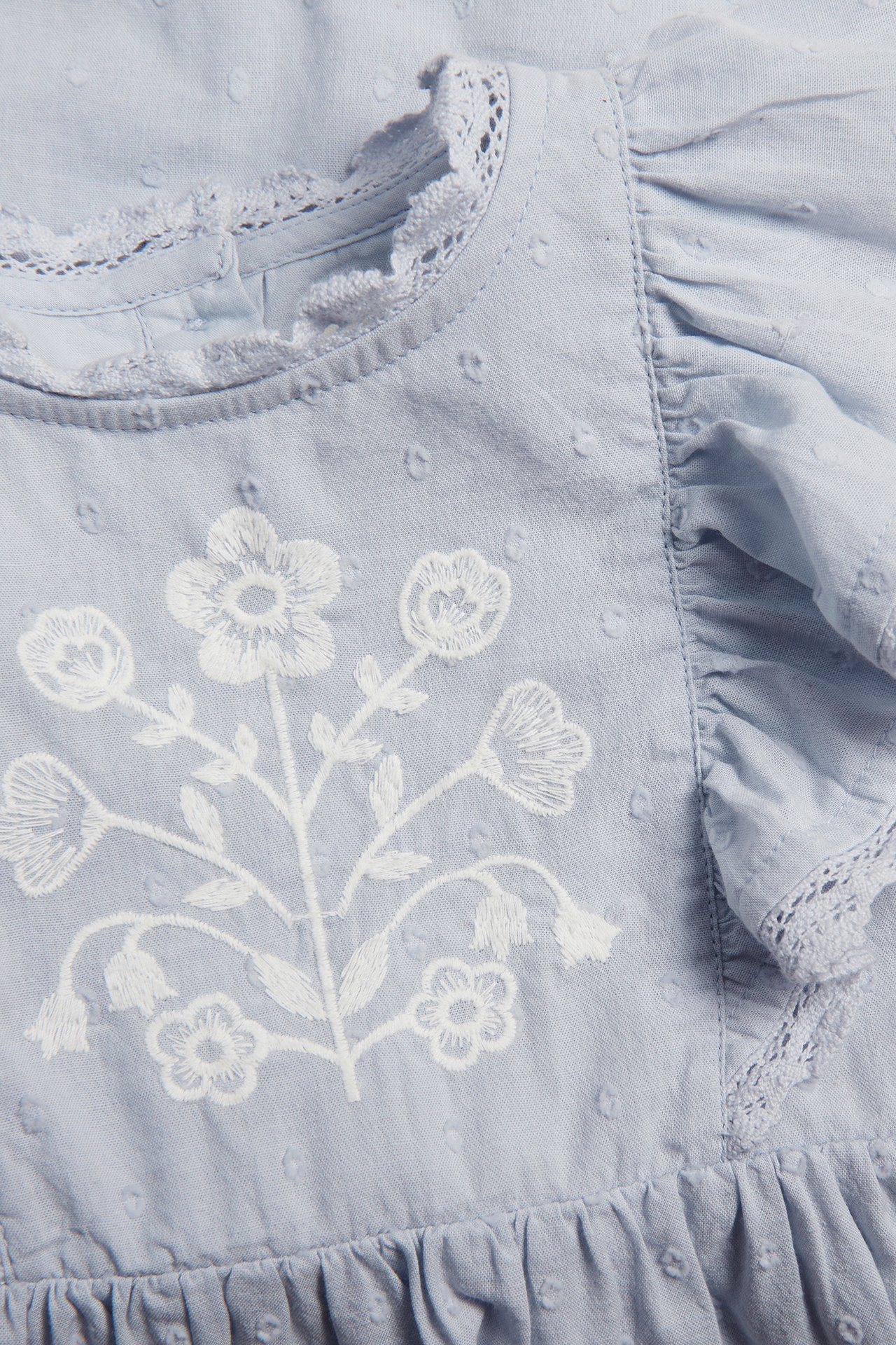 Close-up of a kid's light blue woven cotton dress with white floral embroidery, dot texture, lace trim, and ruffles.