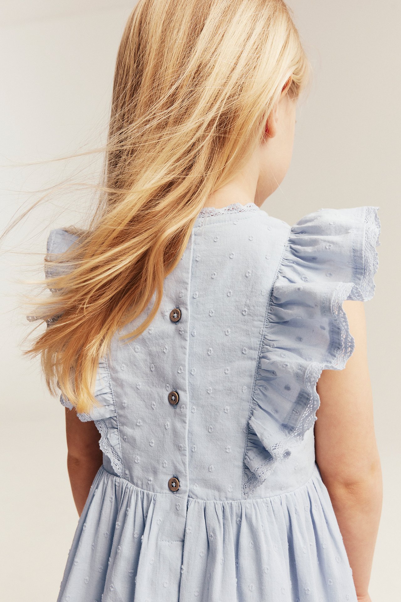 Back view: Kid in a light blue woven cotton dress with dot texture, ruffled lace sleeves, and back buttons.