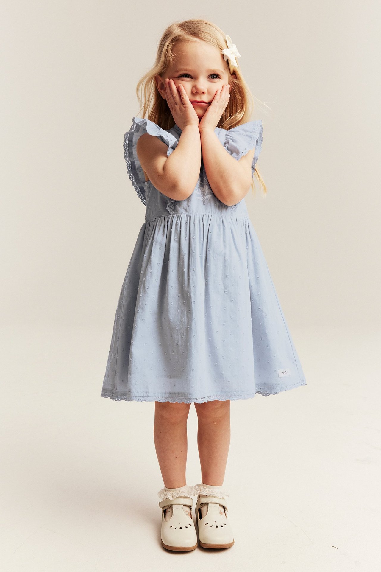 Front view: Kid girl wears a light blue ruffled dress with embroidered flowers, white hair clip, socks, and shoes.