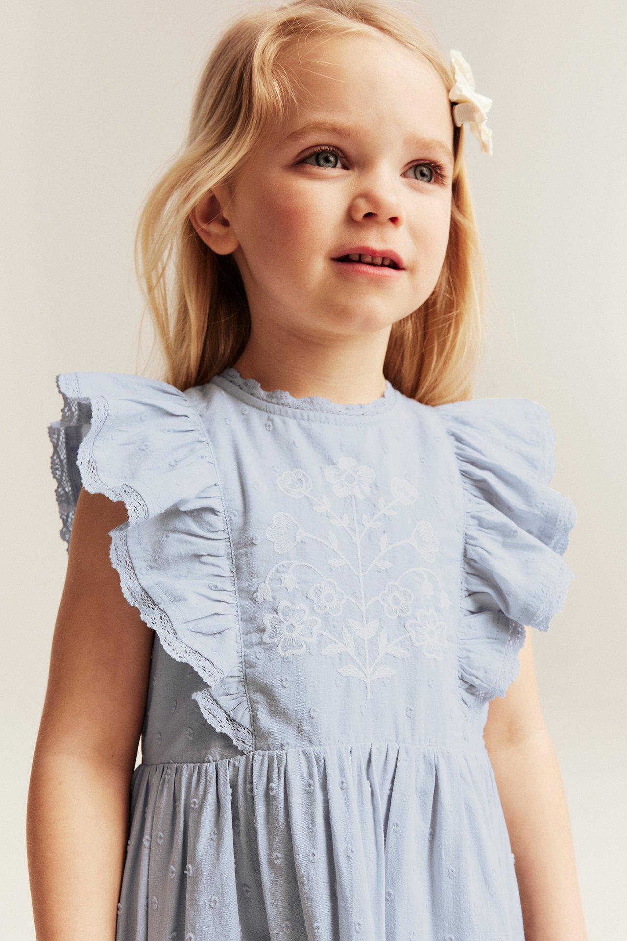 Front view: Kid in light blue dotted cotton dress with white floral embroidery, ruffled sleeves, and hair clip.