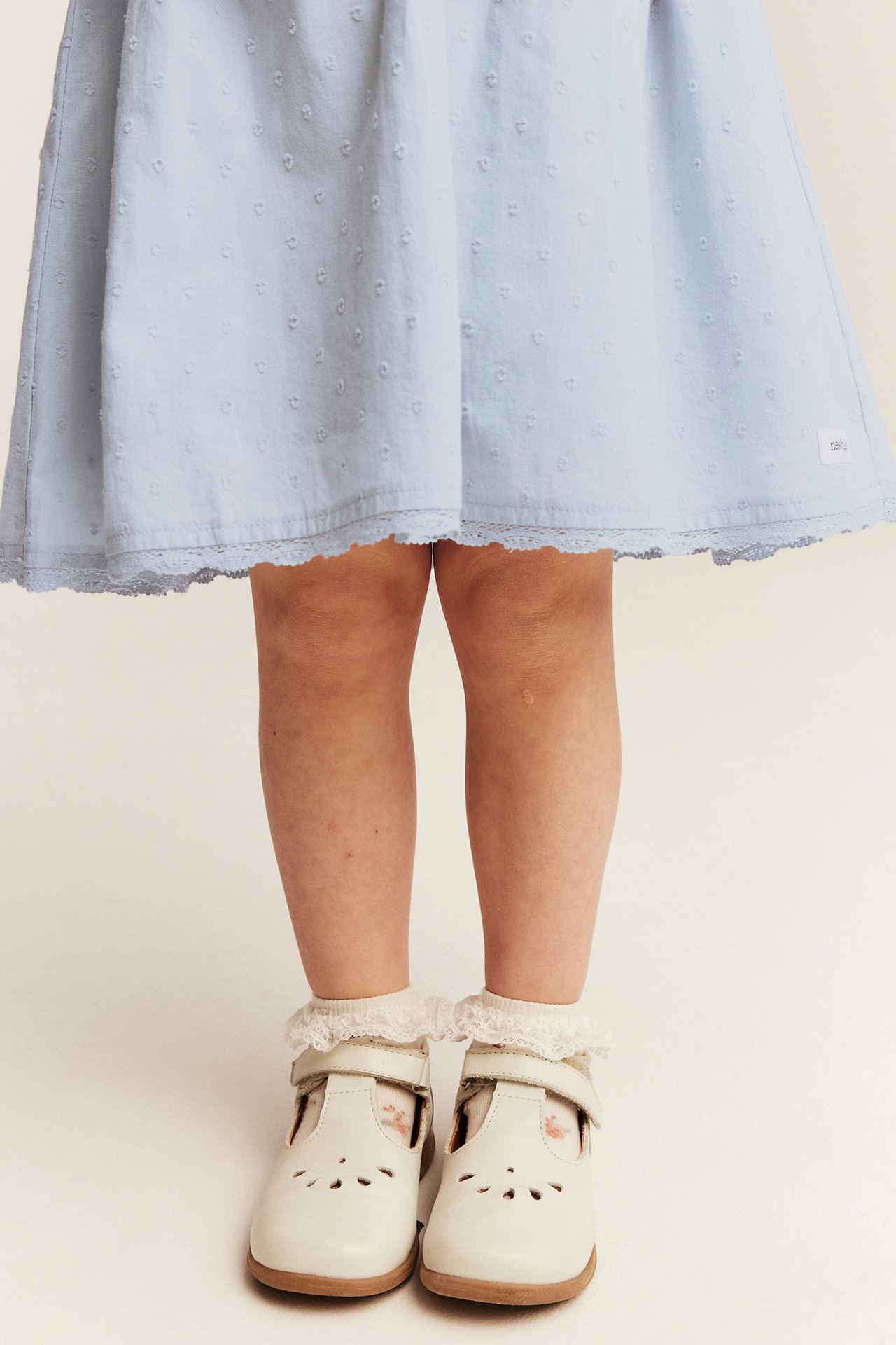 Front view of a kid in a light blue dobby dot cotton dress, white ruffled socks, and white T-bar shoes.