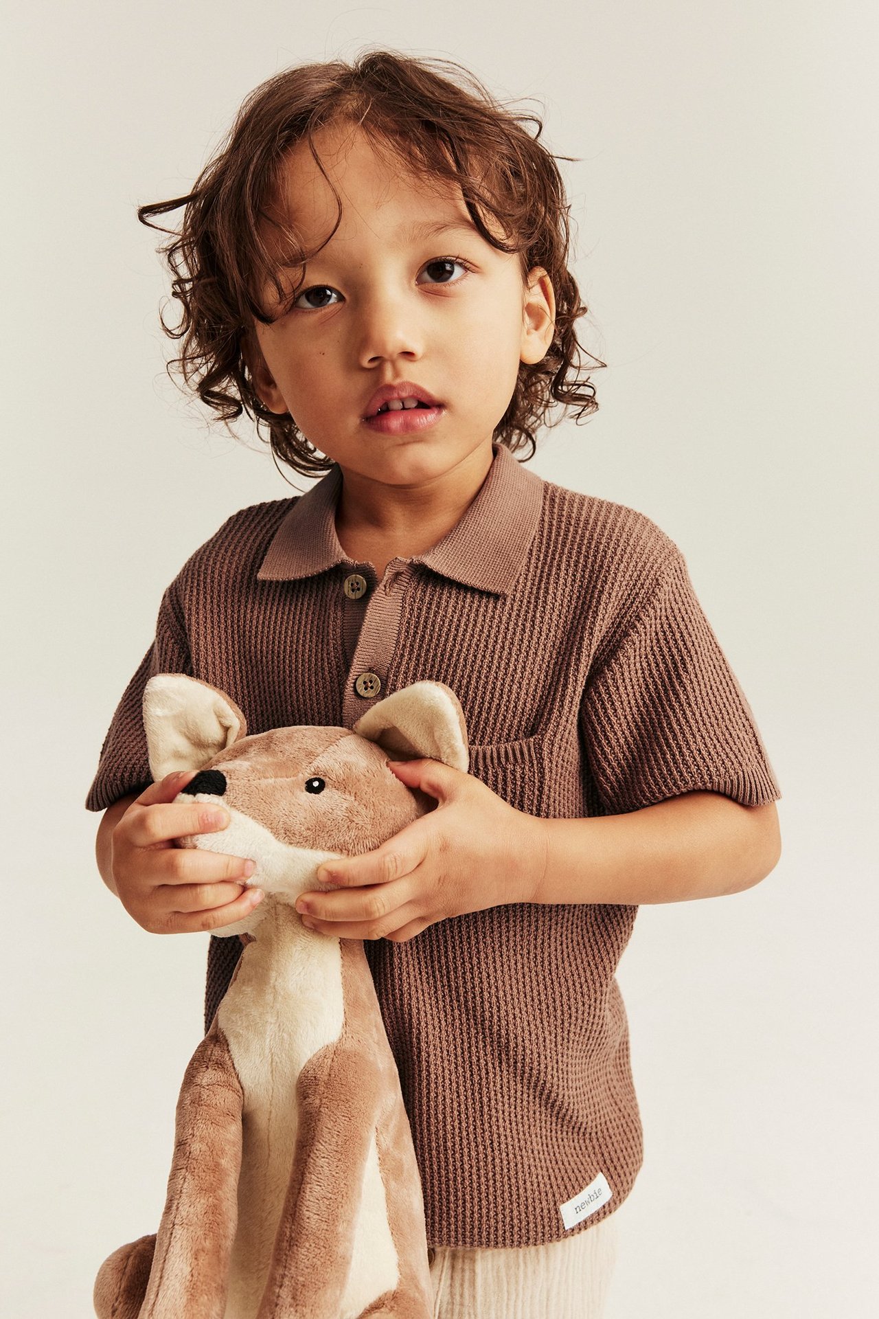 Vorderansicht: Kind in einem braunen, gestrickten Kurzarm-Polohemd mit Tasche, hält ein Plüschtier.