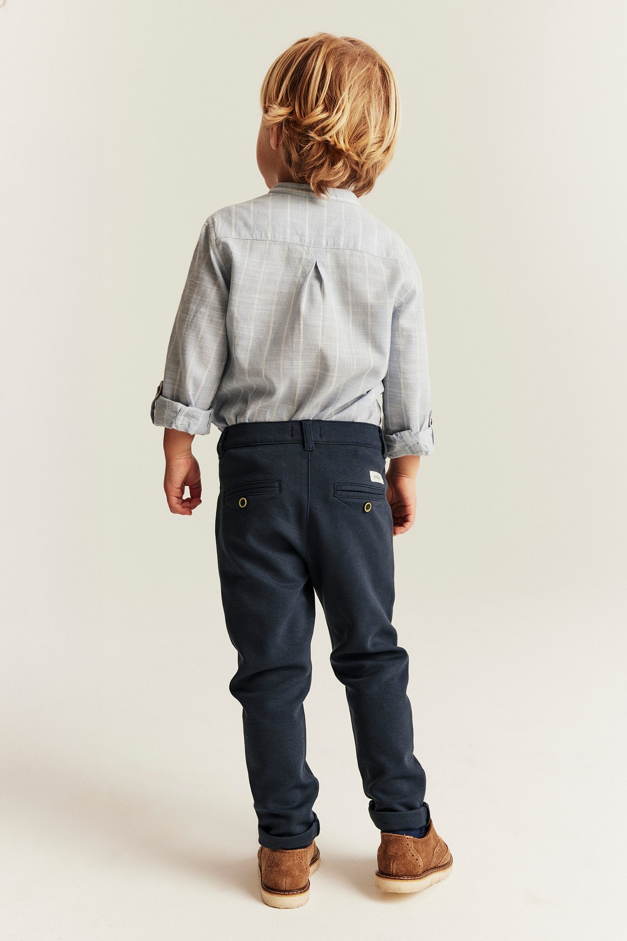 Back view of a kid wearing dark blue trousers with decorative buttons, a light blue striped shirt, and brown shoes.