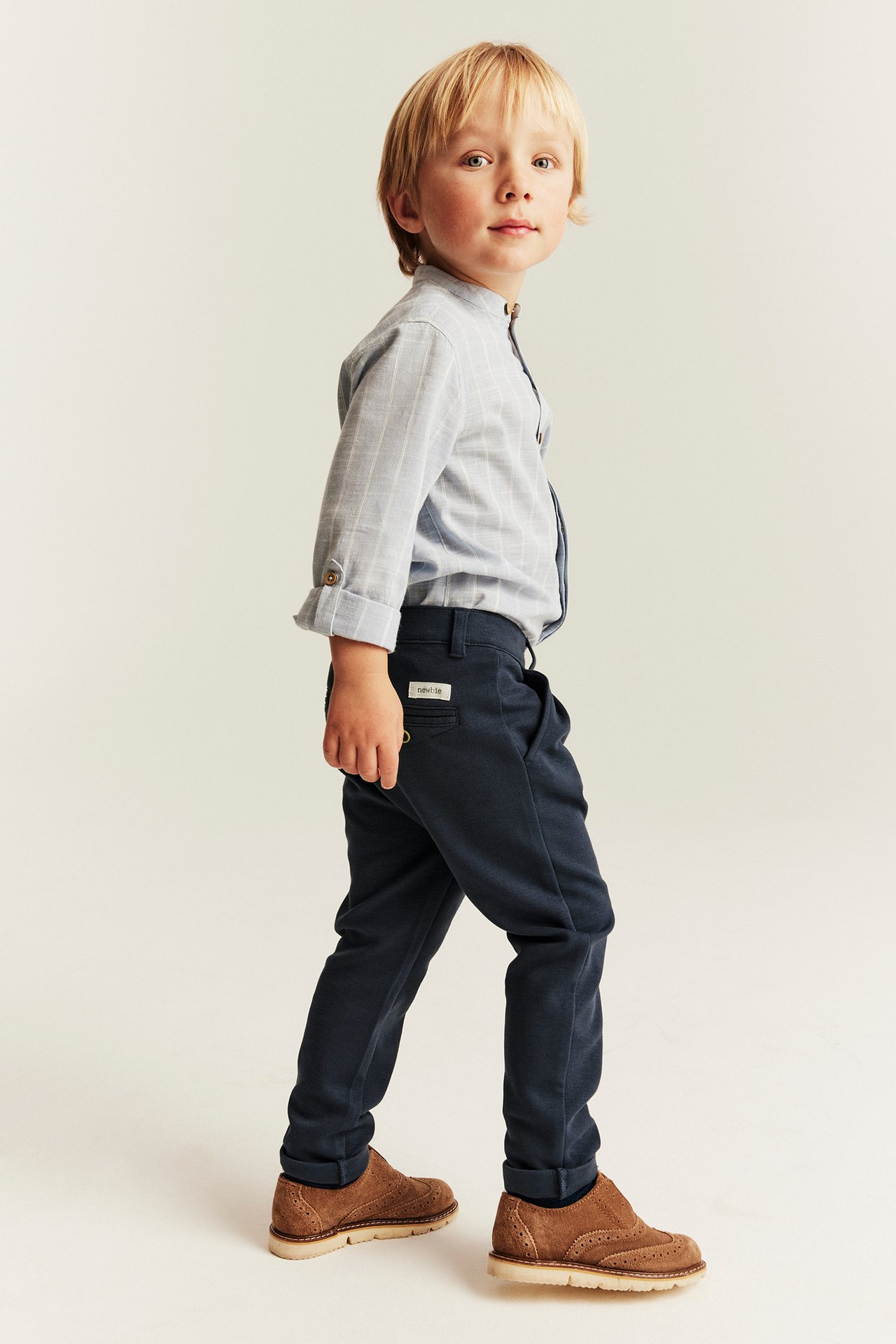 Side view of a kid in dark blue solid pants with cuffed ends, a light blue striped shirt, and brown shoes.