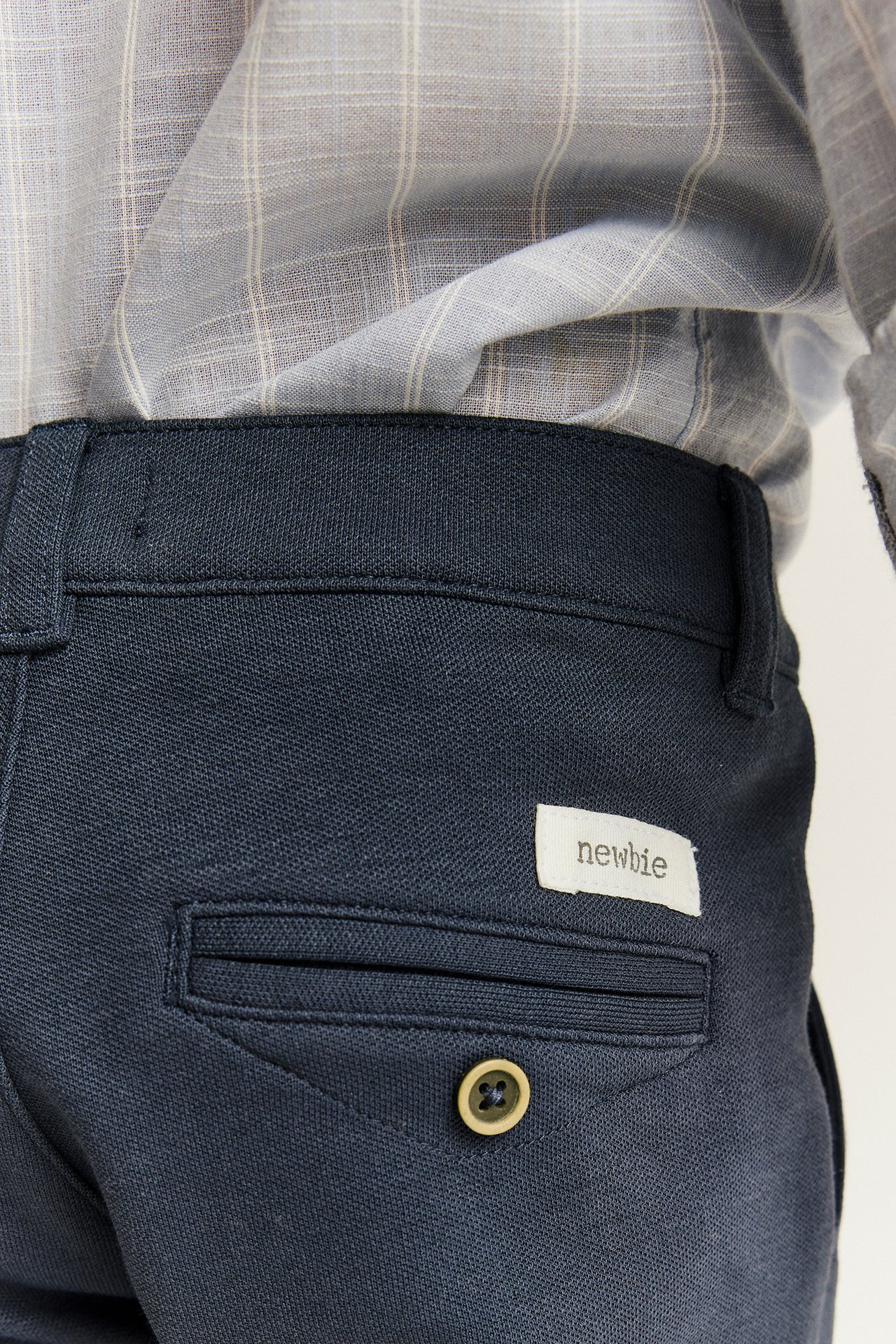 Close-up back view of a kid wearing dark blue trousers with a decorative button and a grey plaid shirt.