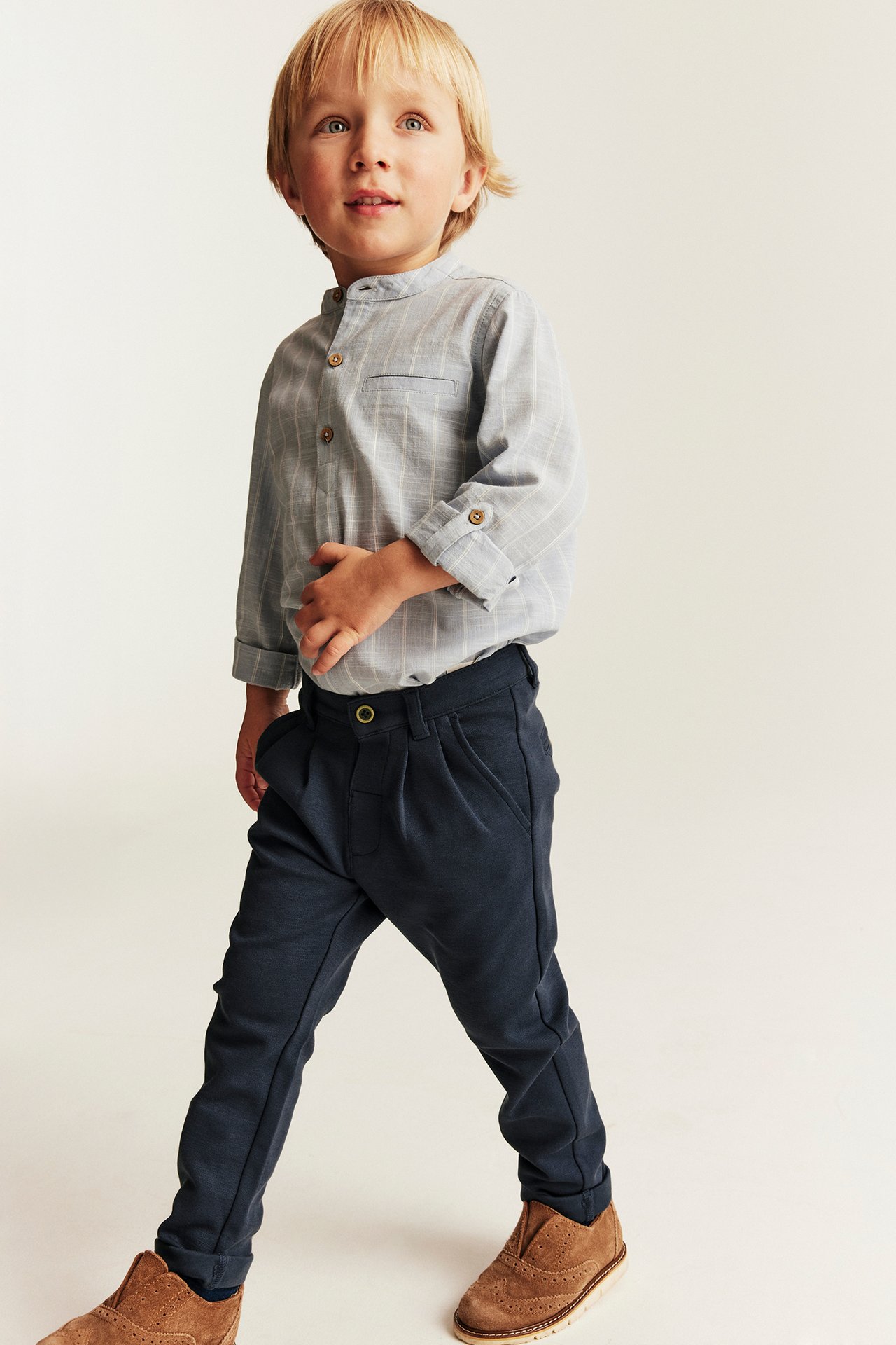 Front-left view: Kid wears dark blue trousers with a button, a striped shirt, and brown shoes.
