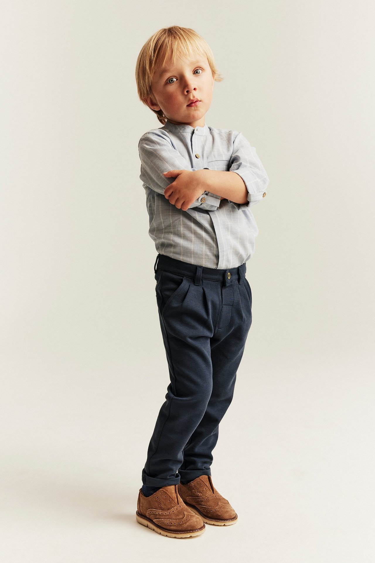Front view of a kid wearing dark blue pleated pants with button, light blue striped shirt, and brown shoes.