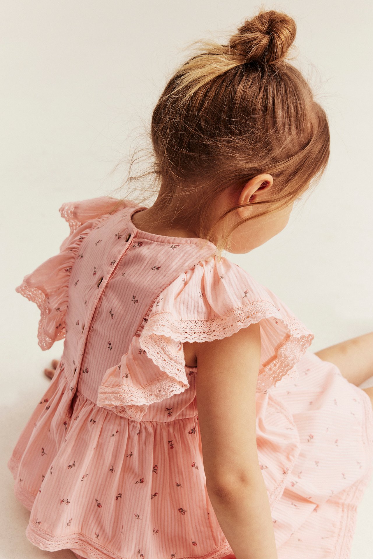Back view: kid in light pink floral striped dress with ruffled, lace-edged sleeves, back buttons.