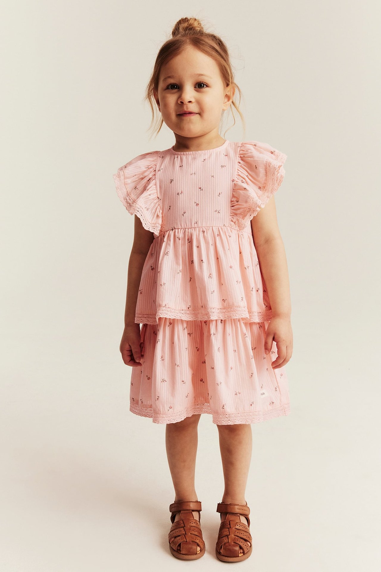 Frontalansicht: Kind trägt ein hellrosa geblümtes Stufenkleid mit Rüschen, Spitze und braunen Sandalen.
