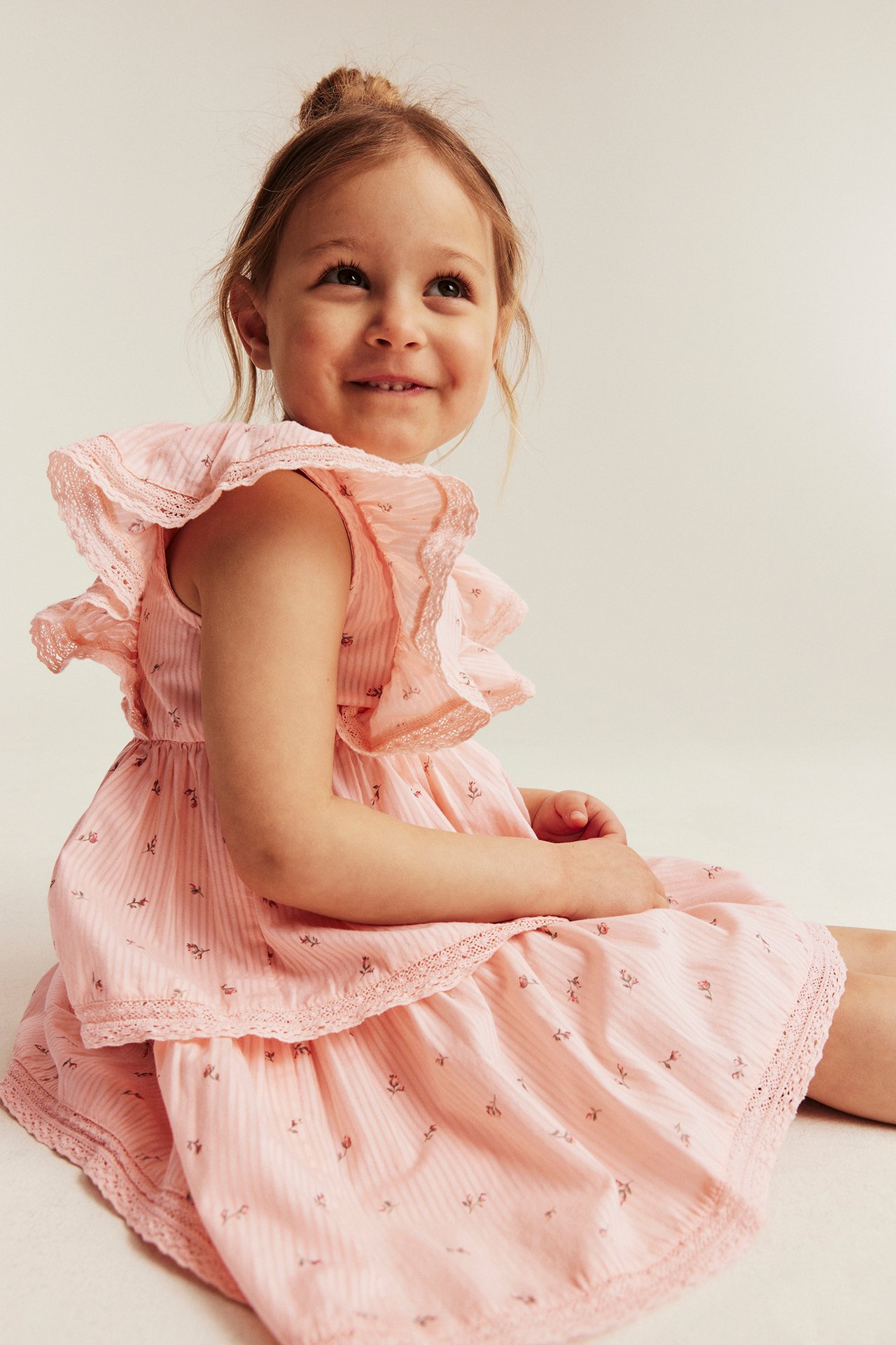 Front-side view of a kid wearing a light pink floral, striped dress with ruffles and lace trim.