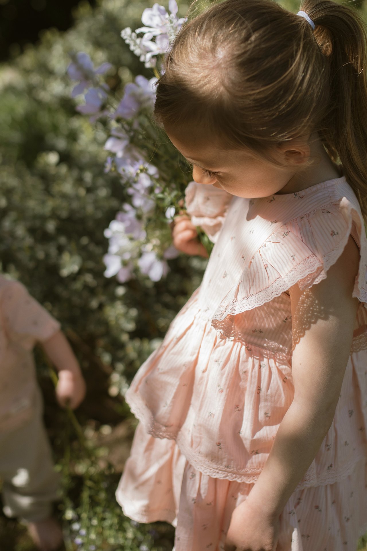 Close-up of kid girl in light pink woven floral dress with tiered ruffles and lace edges, holding a flower.