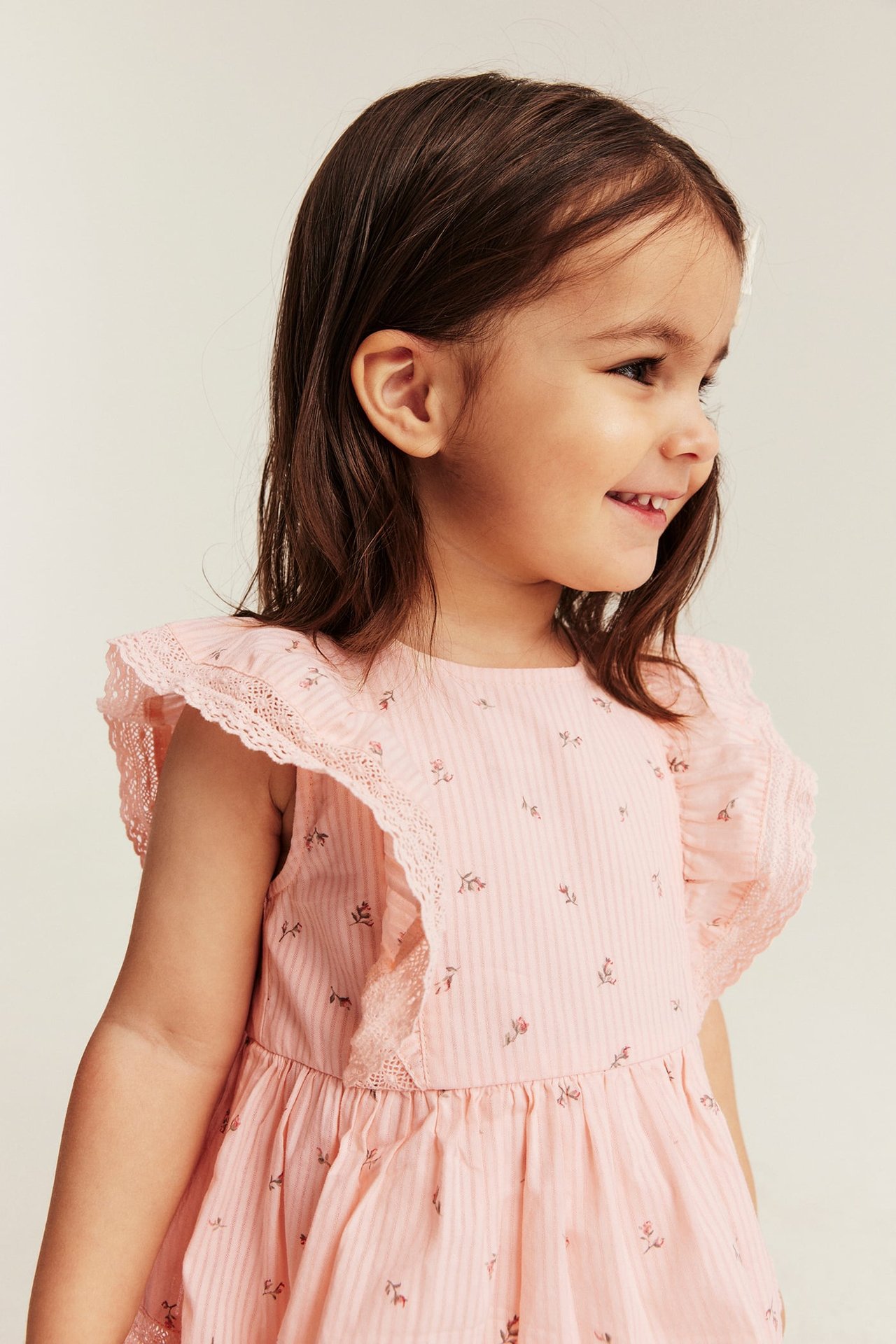 Side view of a smiling kid girl wearing a light pink floral striped dress with lace-edged ruffles.