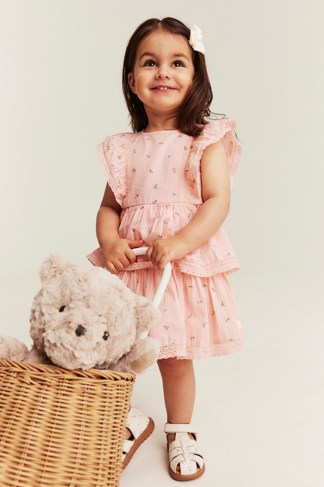 Smiling kid in a light pink floral tiered dress with ruffles, white sandals. Holding a basket and teddy bear.