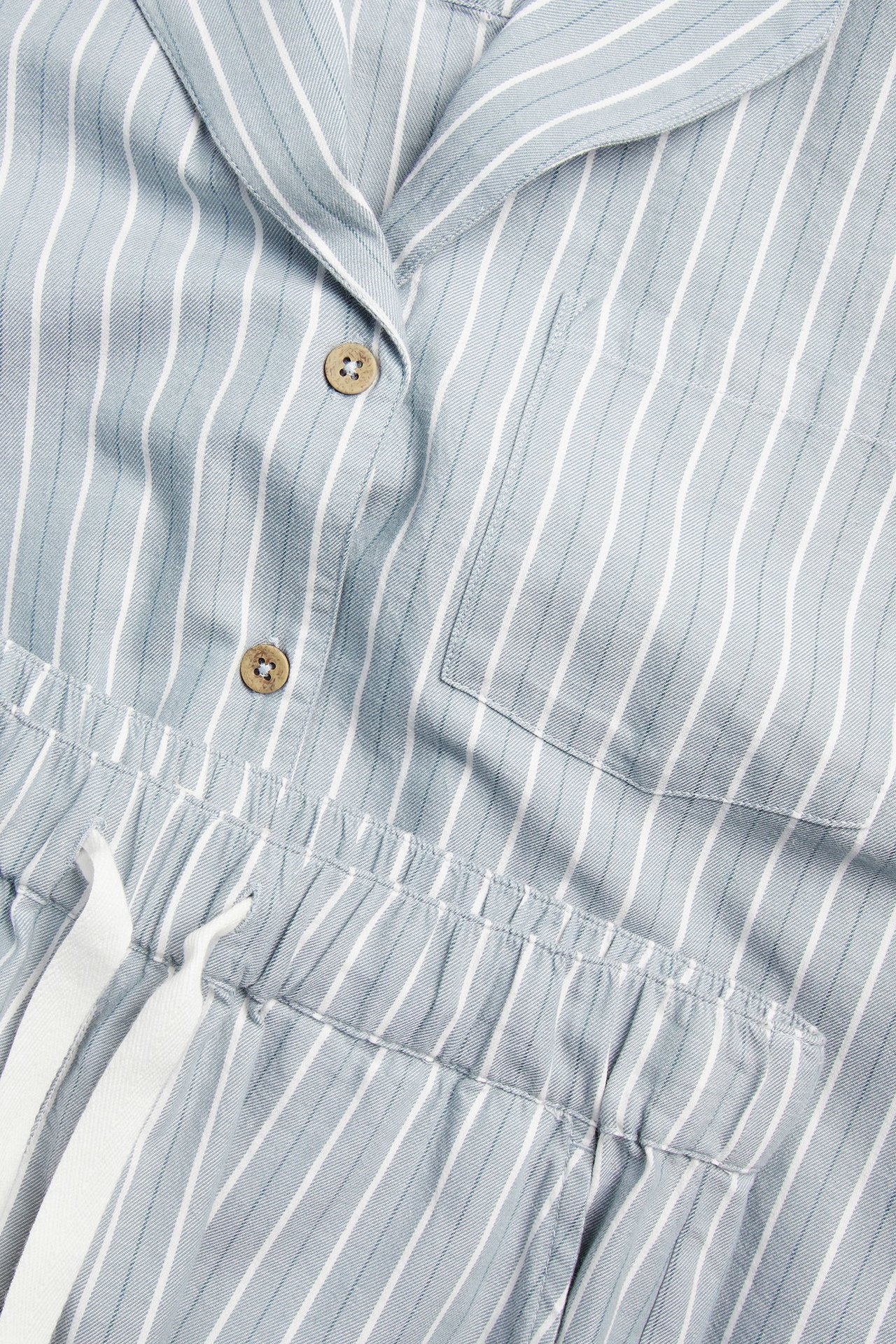 Close-up of woman's light blue and white striped cotton pyjamas. Buttoned collared top with pocket, elasticated shorts with drawstrings.
