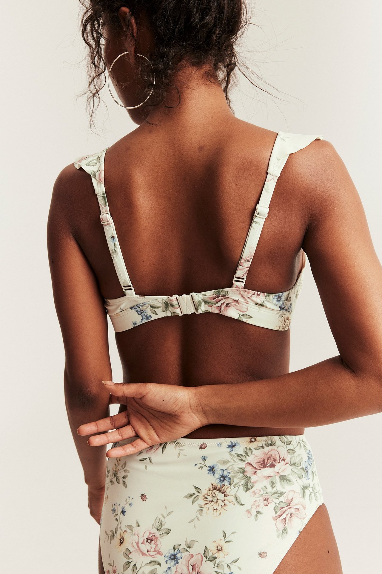 Back view: woman in off-white floral bikini top with ruffled, adjustable straps and matching high-waisted bottoms.