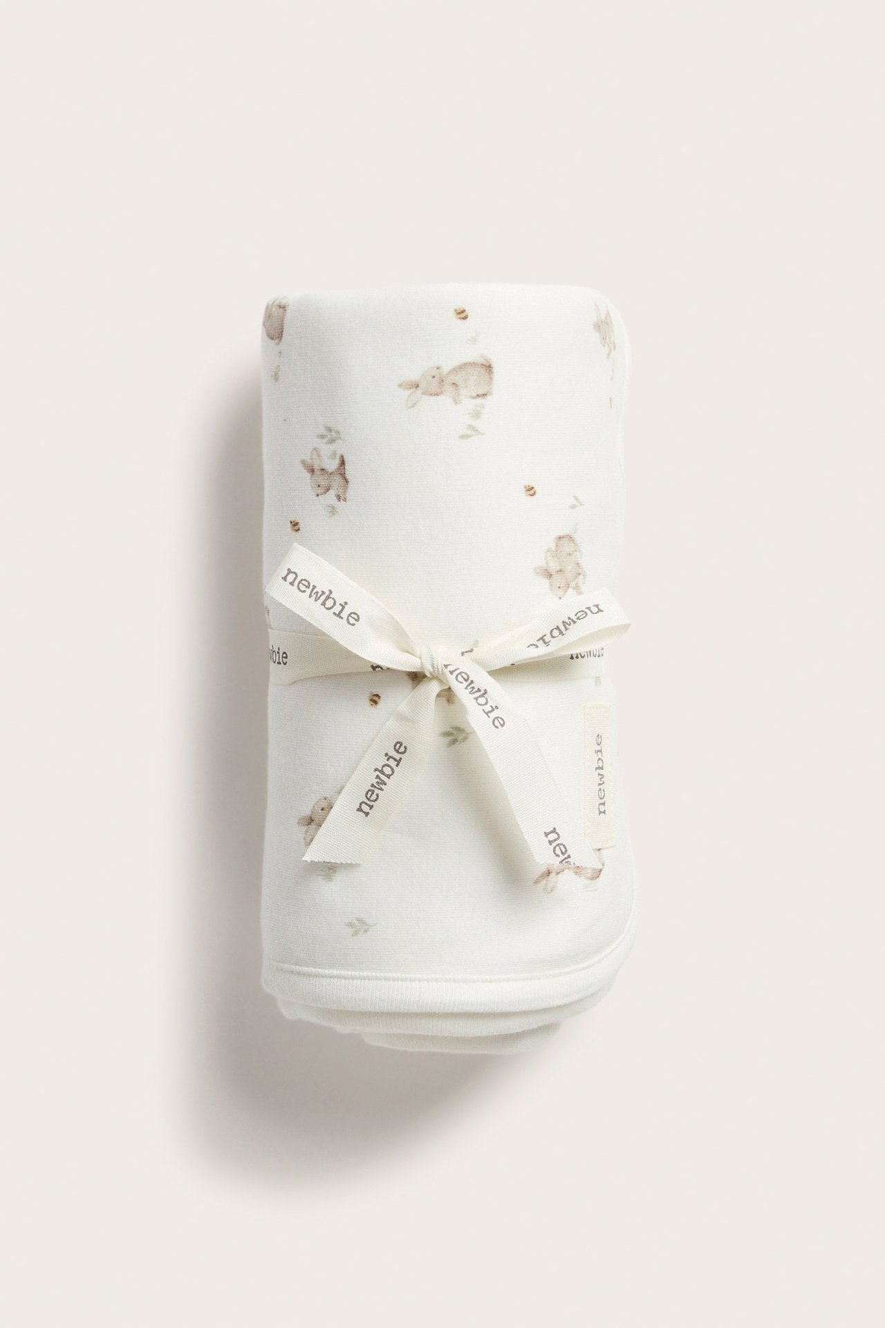 Vorderansicht einer cremeweißen Baumwoll-Babydecke mit Hasenmuster, gerollt und mit einem Newbie-Band gebunden.