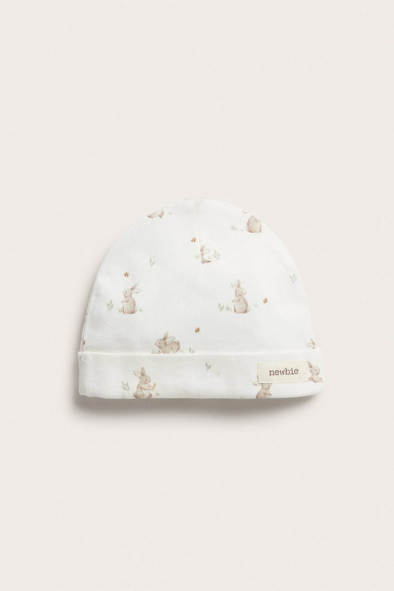 Front view of an off-white baby cotton beanie with a brown bunny pattern and a 'newbie' label.