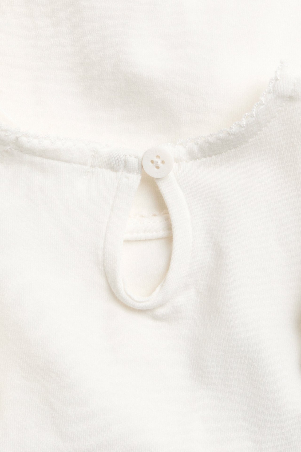 Nahaufnahme: Off-white Babybody aus Baumwolle, rückseitiger Halsausschnitt mit Schlüsselloch, Knopf und Picotstich.