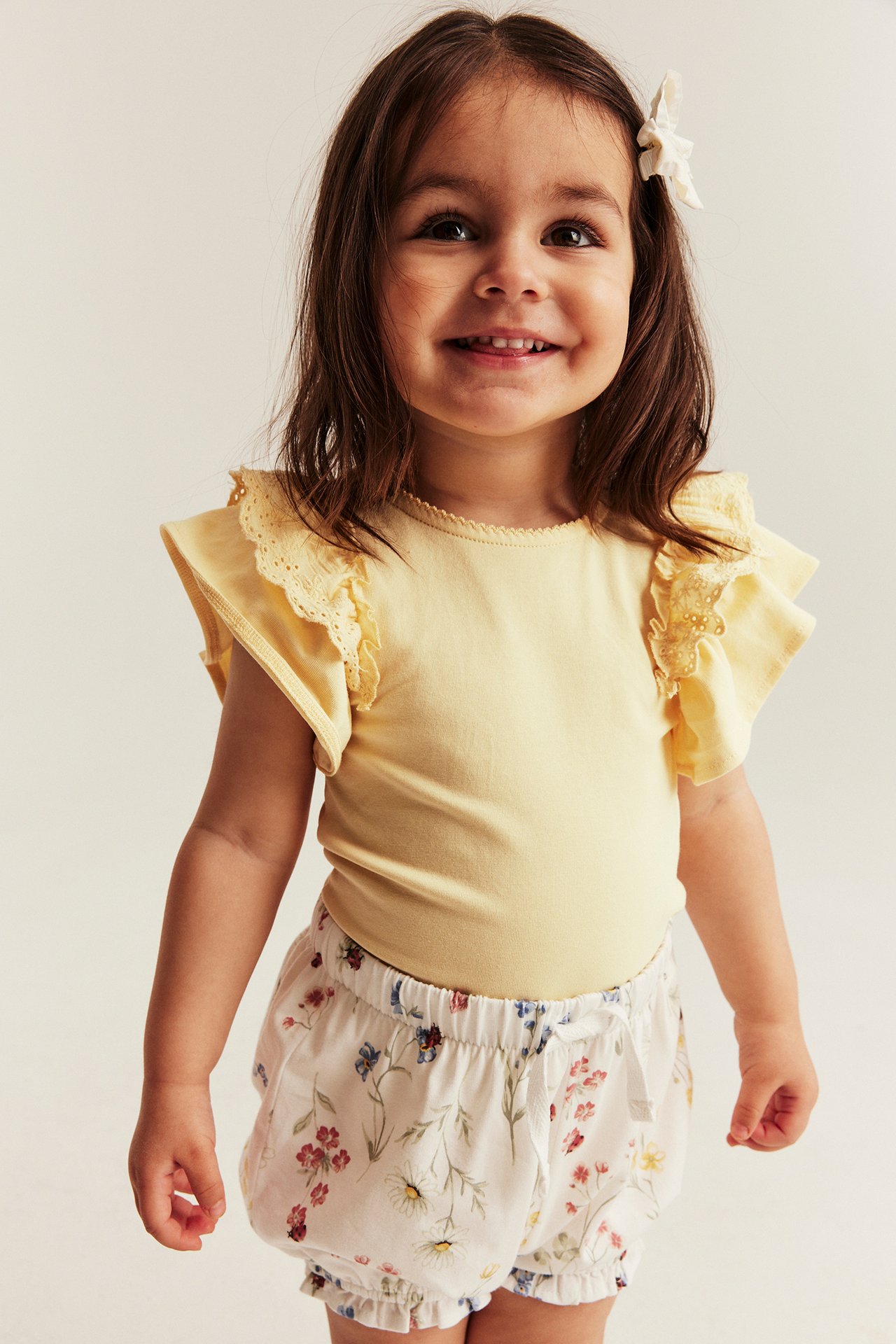 Front view of a smiling kid in a yellow ruffle-sleeved lace bodysuit and white floral shorts.