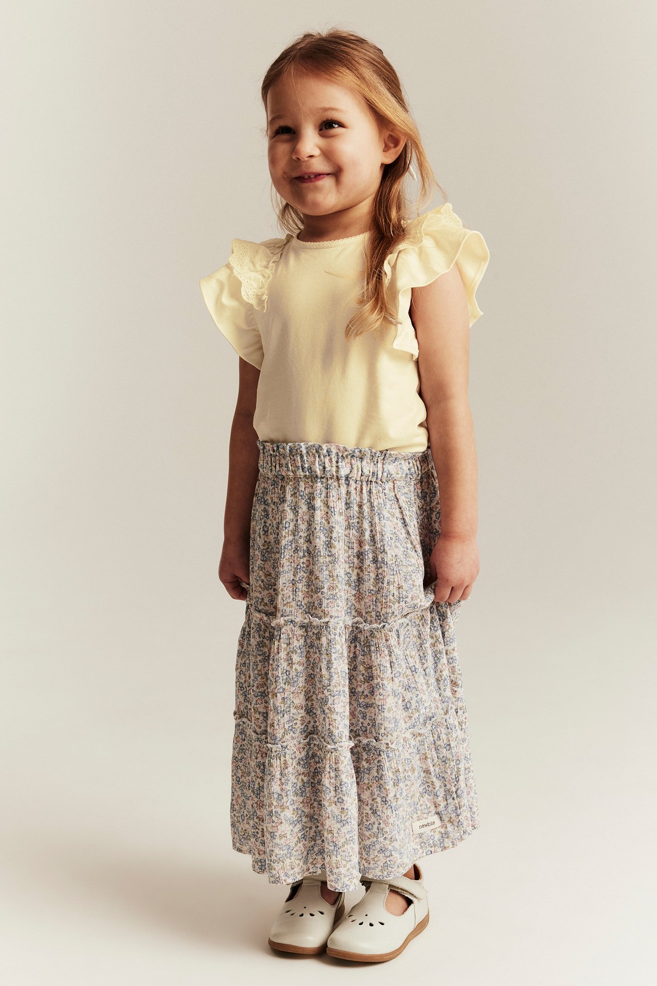 Kid in yellow short-sleeved top with ruffled lace shoulders, a floral skirt, and light T-bar shoes.