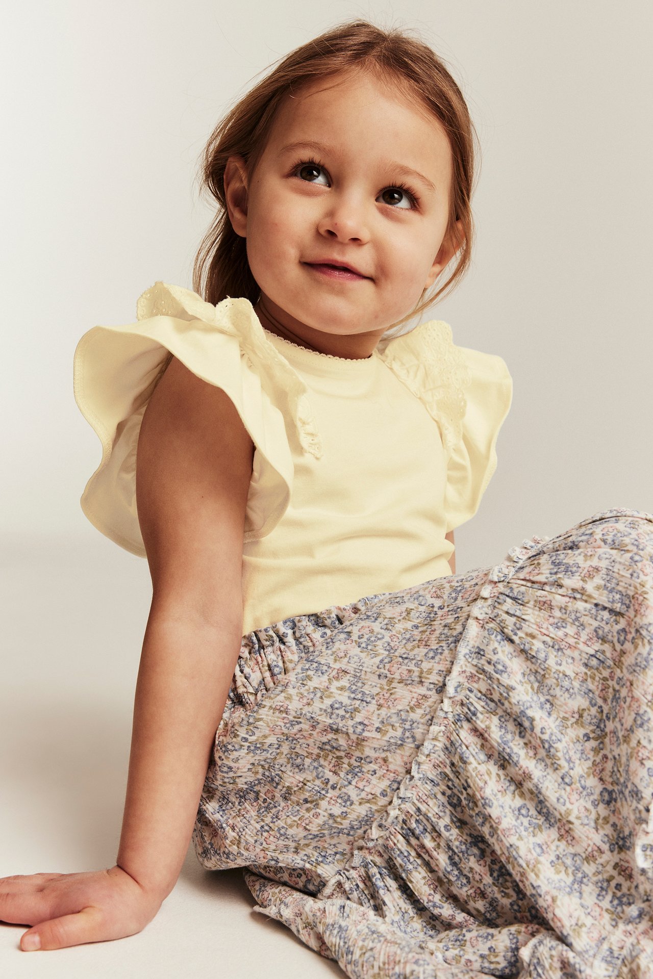 Front view of a kid in a yellow short-sleeved top with lace ruffle sleeves and a floral patterned skirt.
