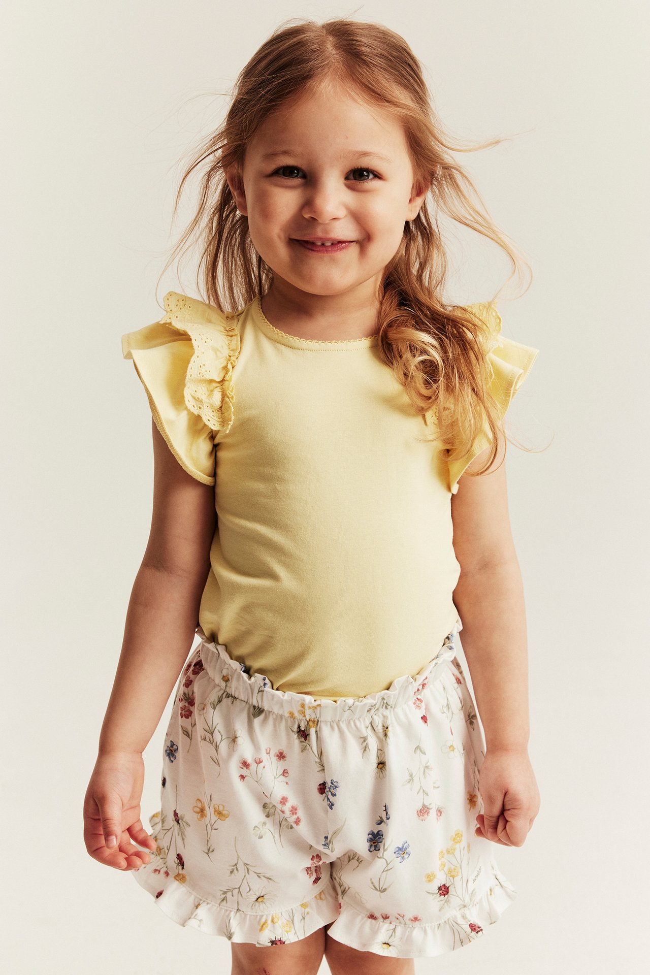 Front view of a kid wearing a yellow short-sleeved top with lace ruffles, and white floral shorts.