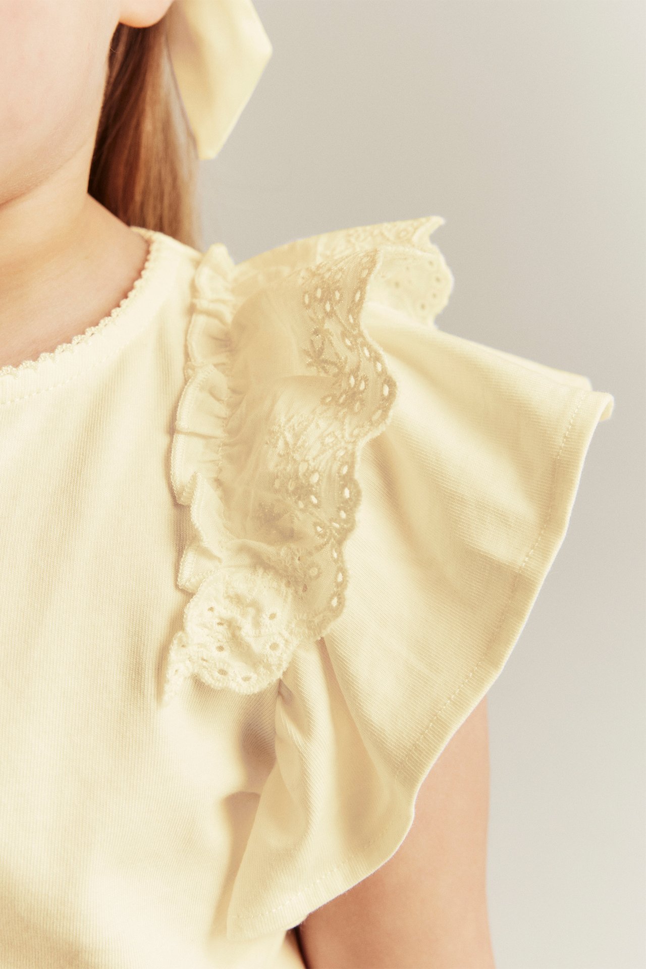 Close-up: Kid wears a light yellow short-sleeved top with ruffle sleeves, lace shoulder, and a hair bow.