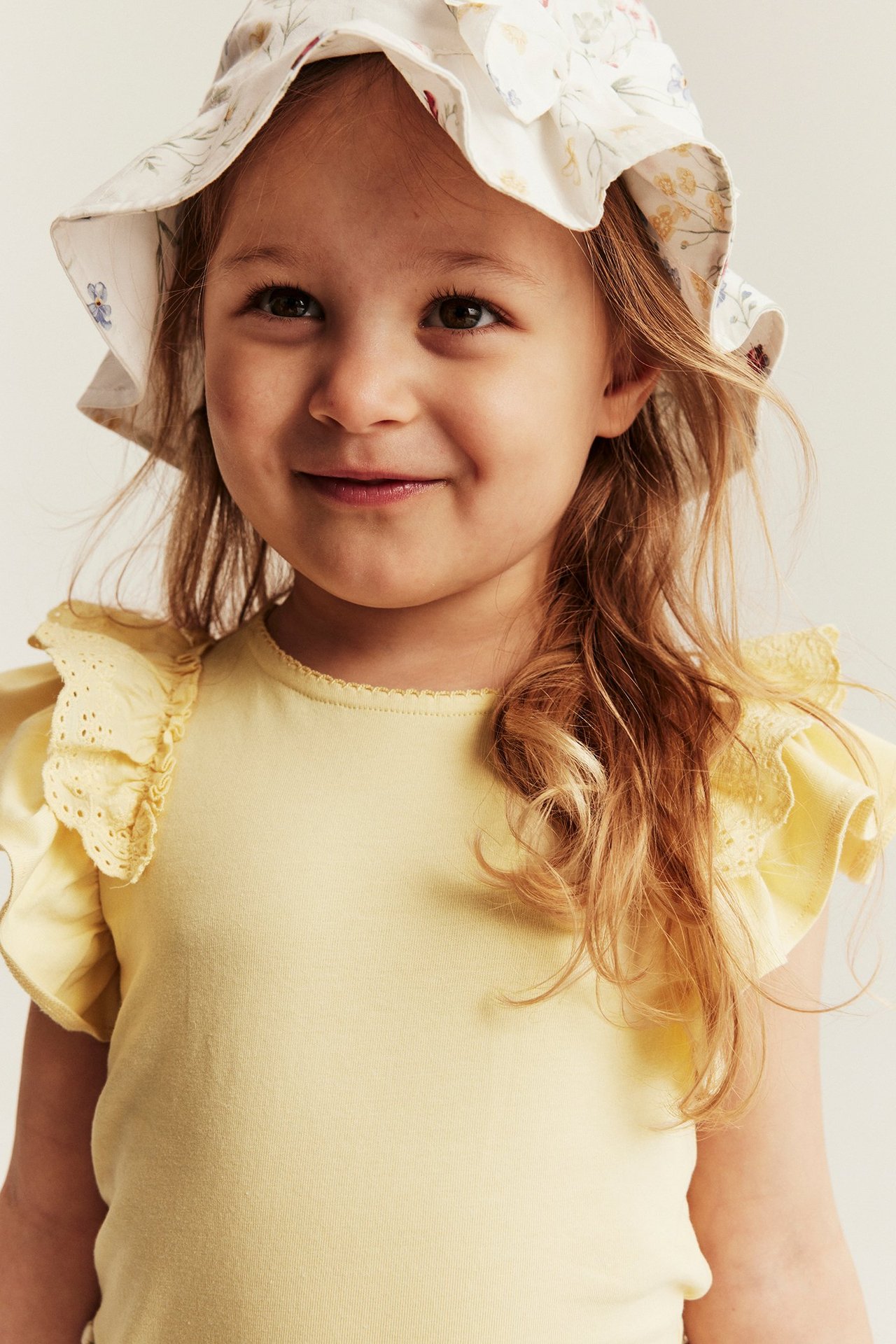 Nahaufnahme, Vorderansicht eines Kindes in gelbem Kurzarm-Top mit Spitzenrüschen und floralem Fischerhut.