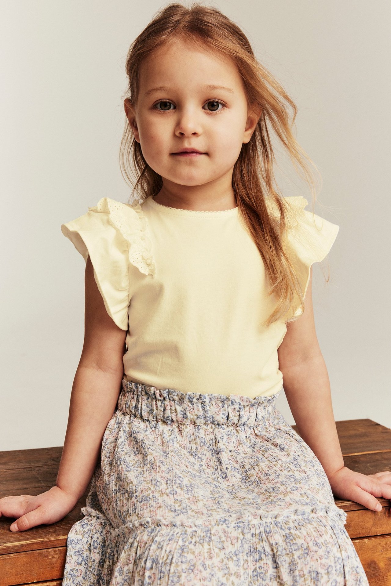 Vorderansicht eines Kindes in gelbem kurzärmeligem Top mit Spitzenrüschen und floral gemustertem Rock.