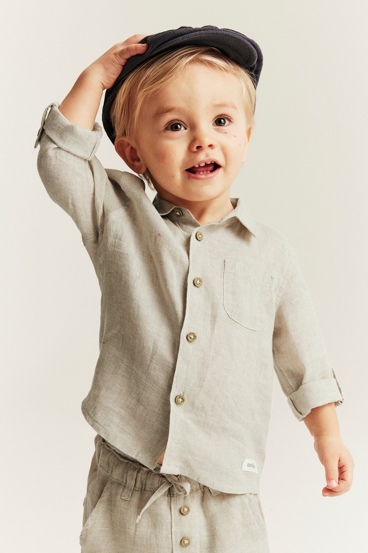 Front view of a kid wearing a light beige linen collared shirt with rolled-up sleeves and dark blue cap.