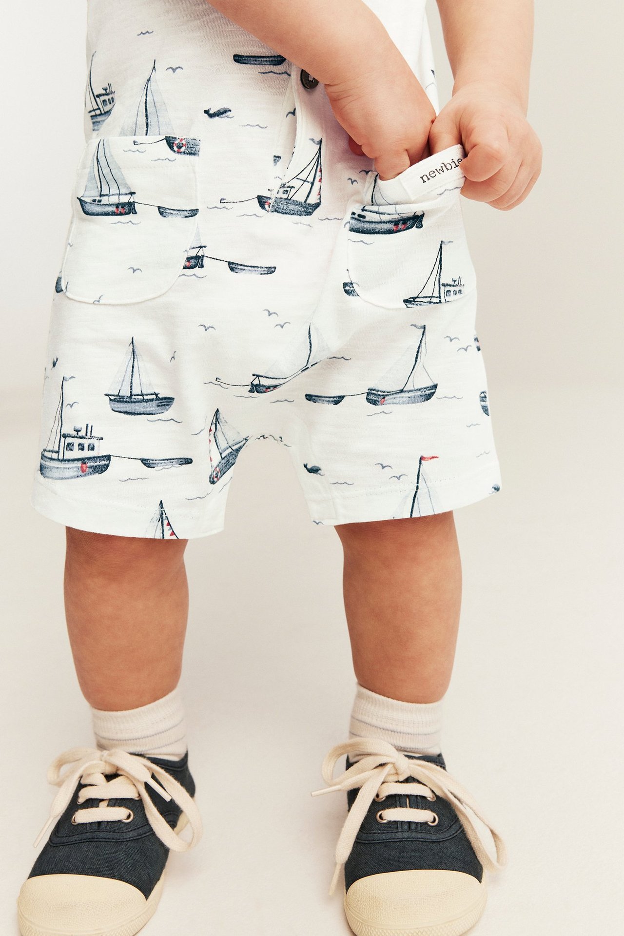Front view: baby in off-white sailing boat print slub jersey romper with patch pockets and dark blue shoes.