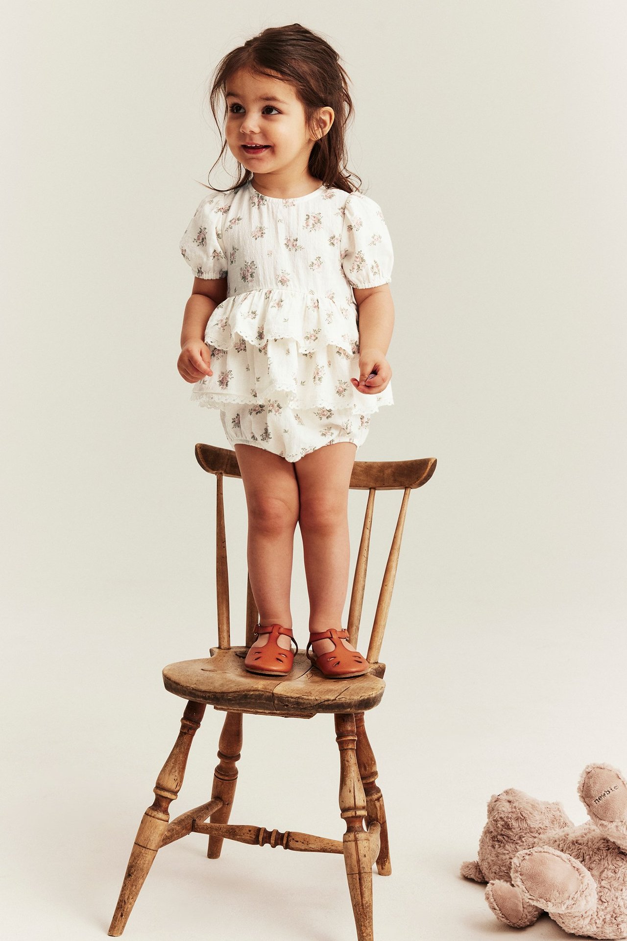 Vorderansicht eines Kindes in einem cremeweißen floralen Baumwoll-Overall mit Rüschen und braunen Sandalen.