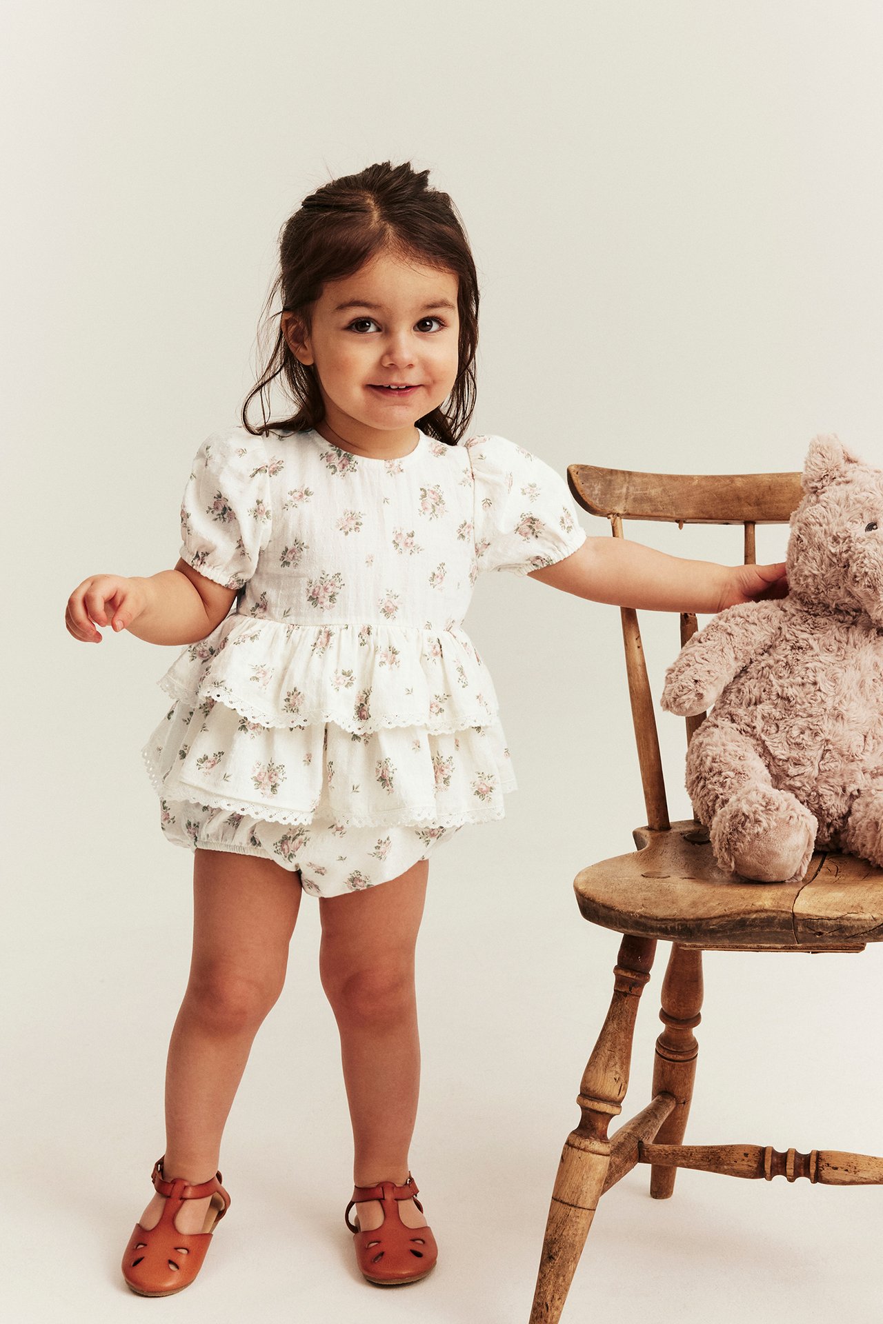 Front view: Kid wears an off-white floral ruffled jumpsuit with puffed sleeves, brown sandals, touching a teddy bear.