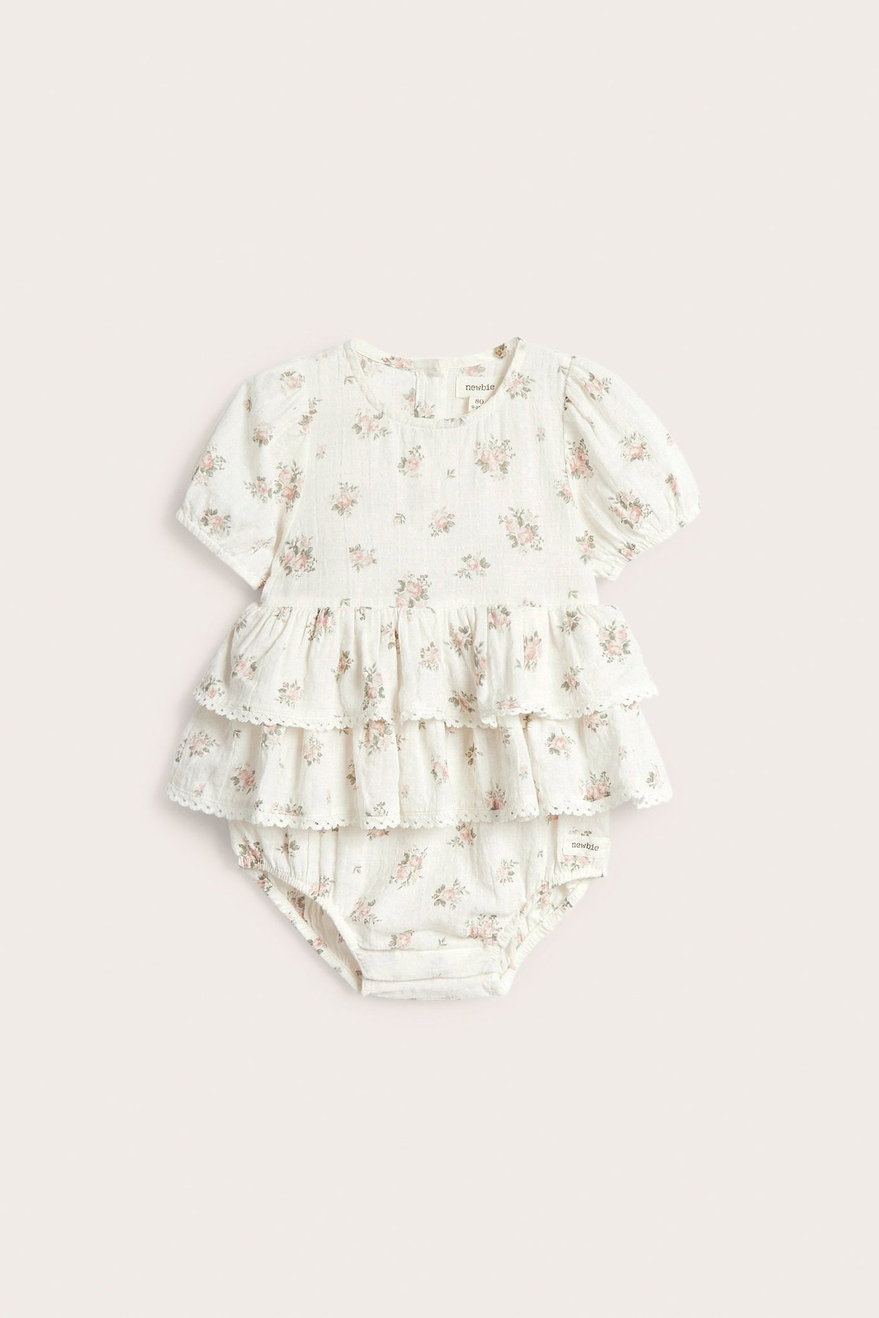 Vorderansicht eines cremeweißen Baby-Baumwoll-Overalls mit Blumenmuster, Puffärmeln und Stufenrüschen.