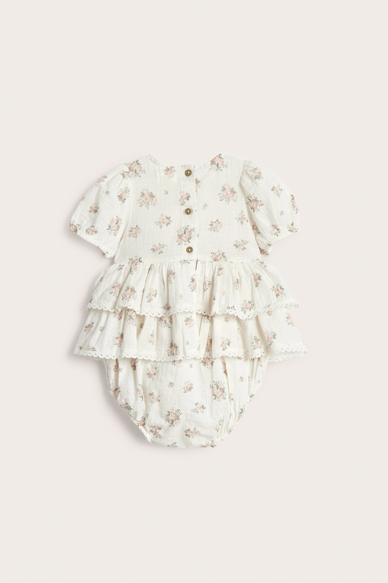 Rückenansicht eines cremefarbenen Baby-Baumwoll-Overalls mit Blumen, gestuften Rüschen, Spitze und Knöpfen.