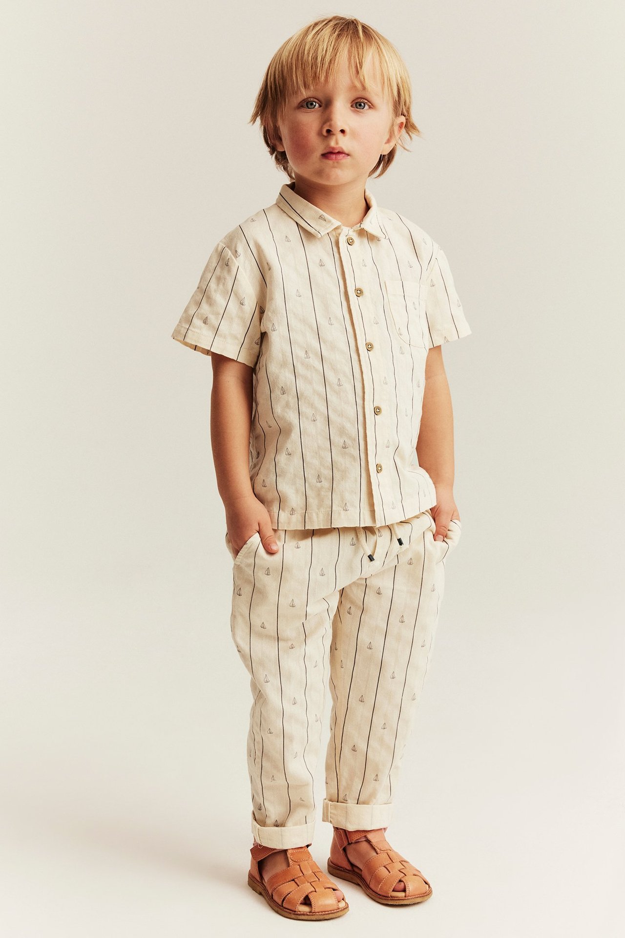 Vorderansicht: Junge in einem hellbeigen gewebten Kurzarmhemd mit Segelboot-Streifen, passender Hose, braunen Sandalen.