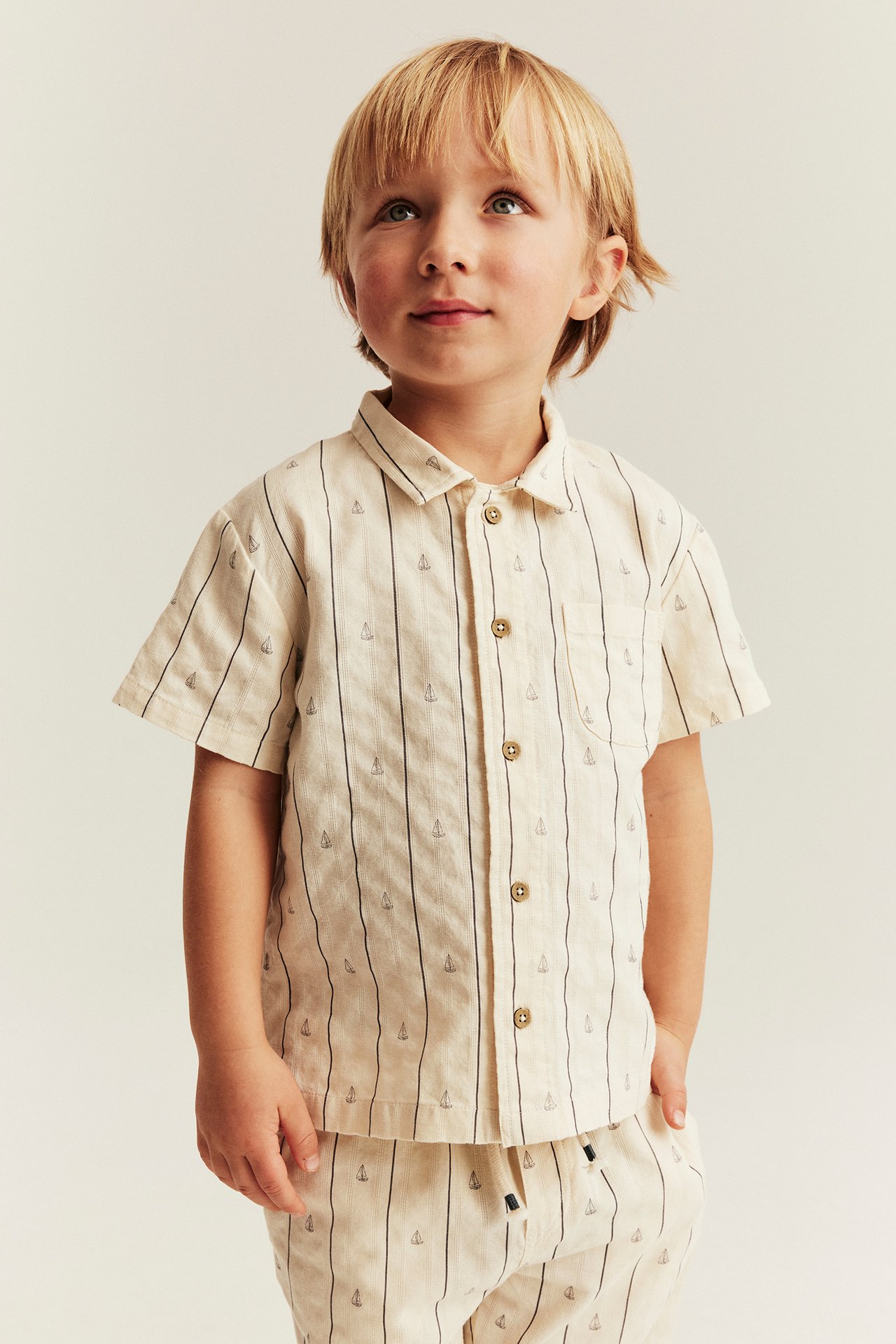 Vorderansicht: Kind trägt ein cremefarbenes gestreiftes kurzärmeliges Baumwollhemd mit Segelbooten und passende Hose.