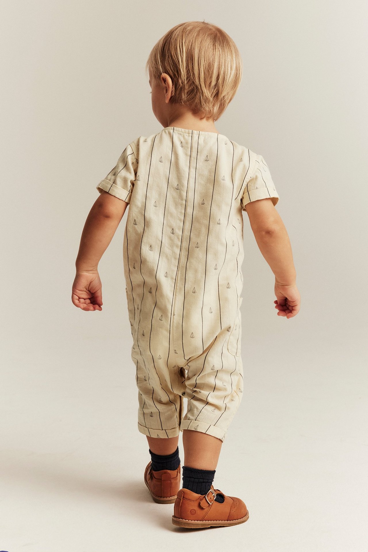 Back view of a baby in an off-white striped cotton jumpsuit with sailboats, dark socks, and brown shoes.