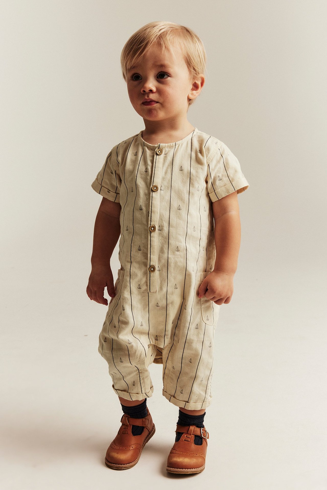 Front view: baby in a white cotton short-sleeved jumpsuit with navy stripes and sailboats, brown shoes, dark socks.