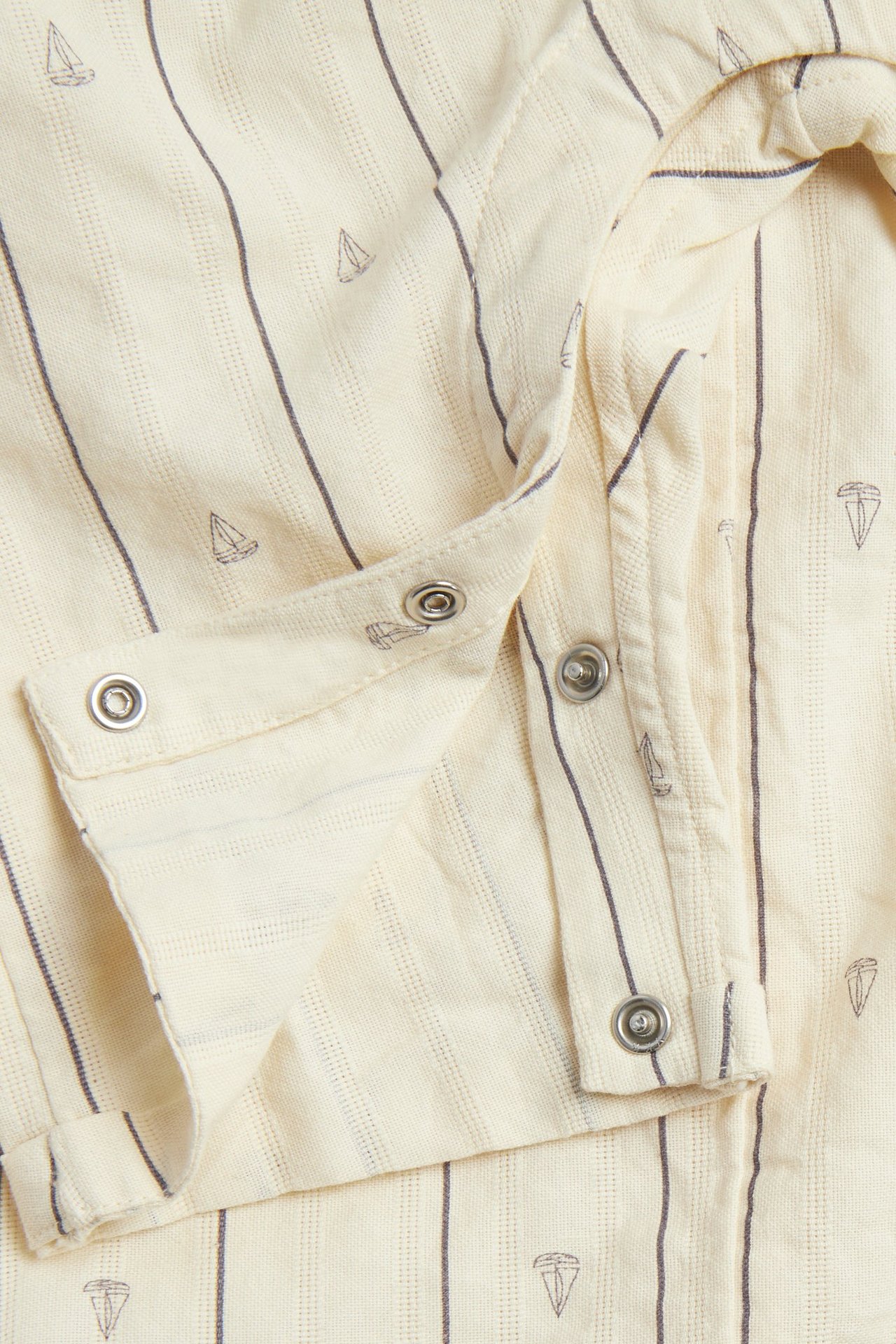 Close-up: white baby jumpsuit with vertical stripes, sailboat print, and silver snap buttons.