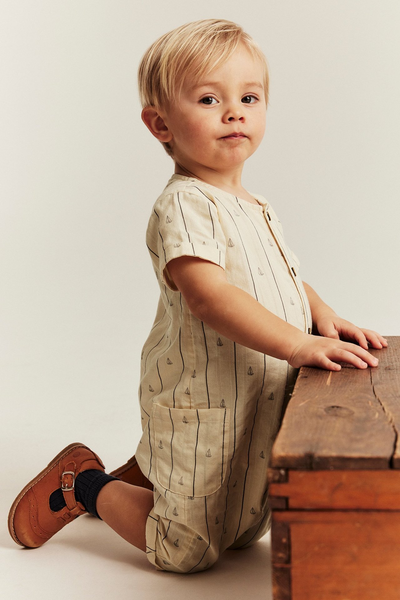 Seitenansicht: Baby trägt einen hellbeigen kurzärmeligen Overall mit Segelboot-Streifen, dunkle Socken und braune Schuhe.