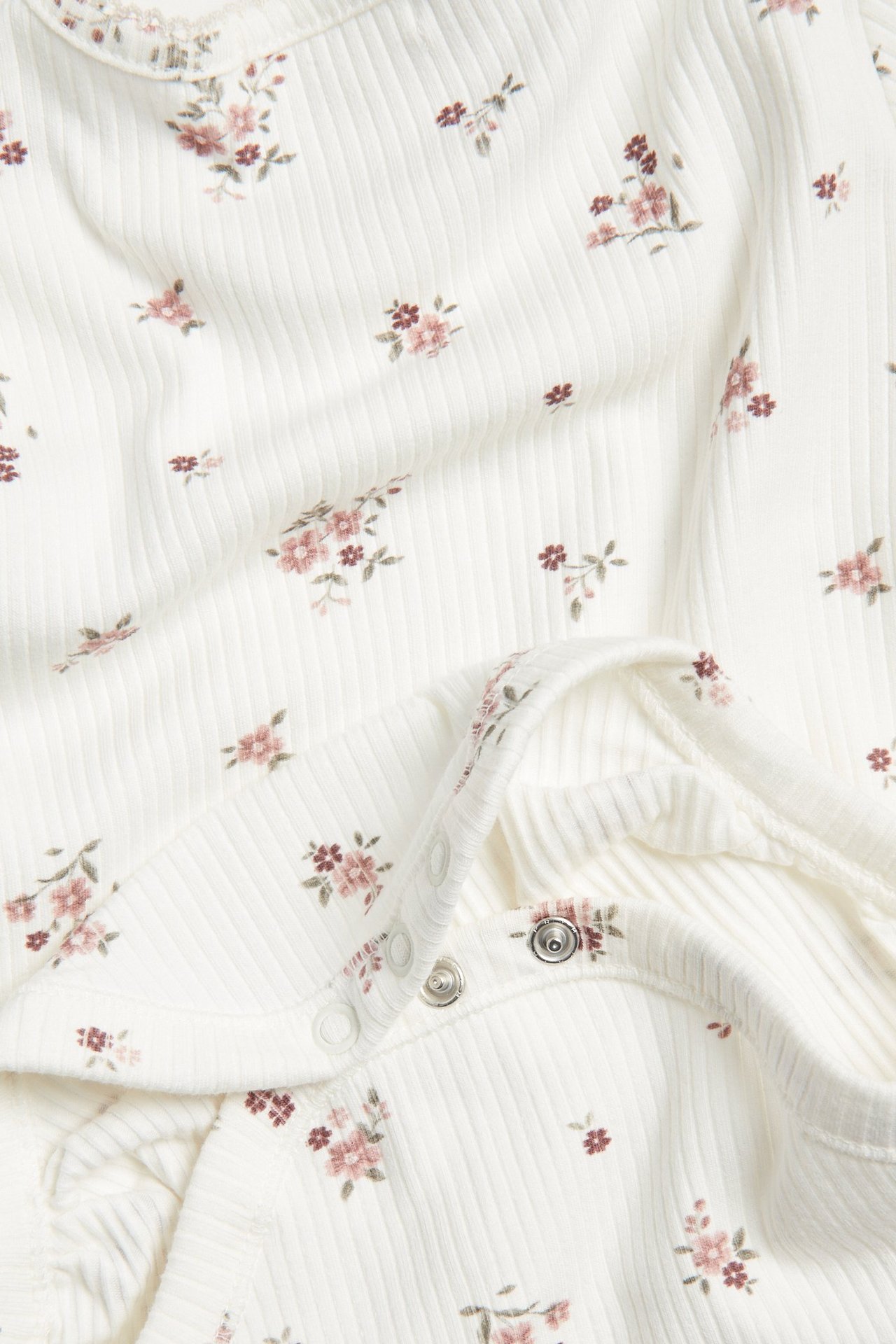 Nahaufnahme eines babyweißen gerippten Bodys mit Blumenmuster und Druckknöpfen an der Schulter.