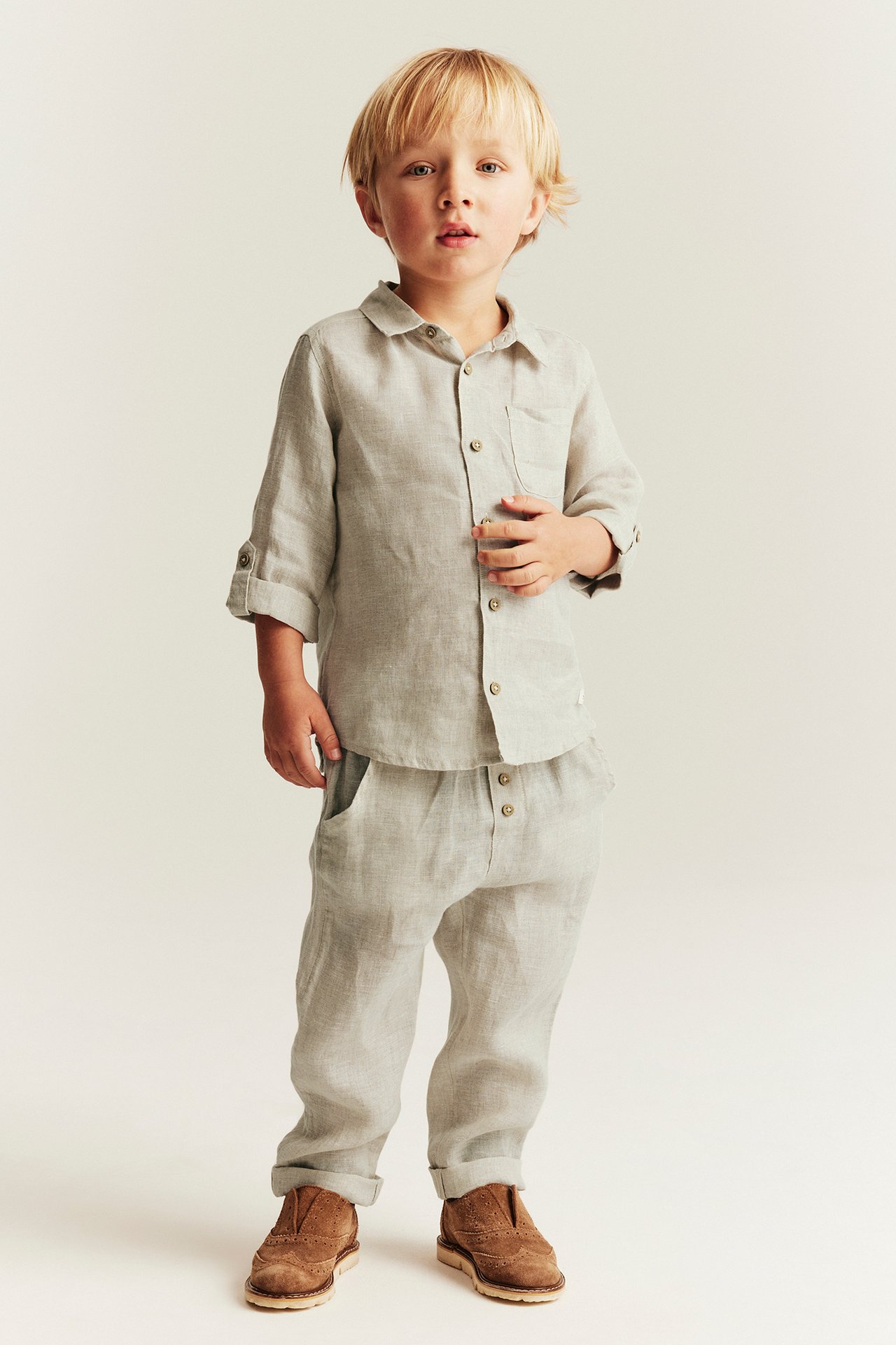 Front view: Kid boy in light beige linen trousers with button fly, matching button-up shirt, and brown shoes.