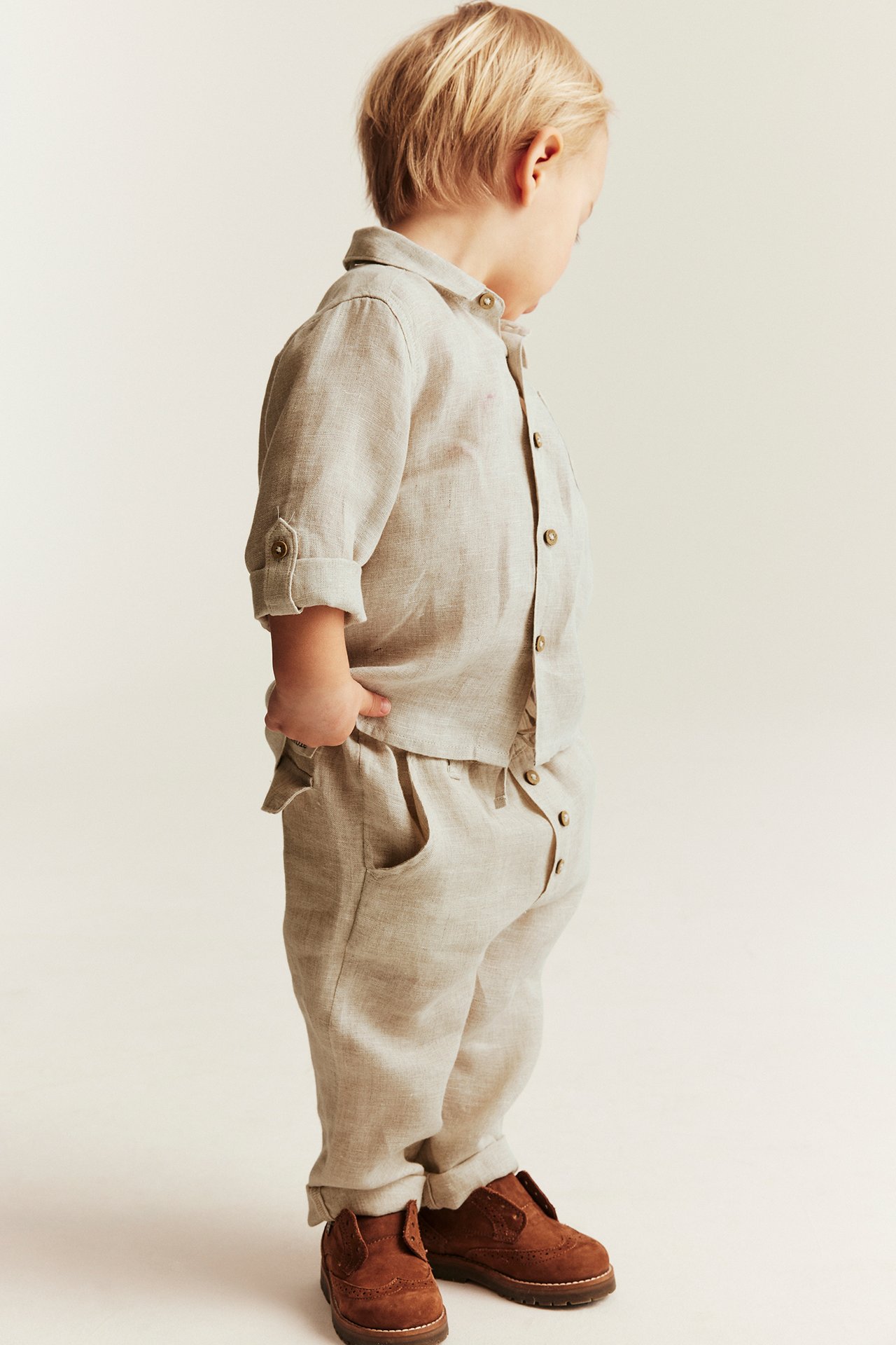 Side view of a kid wearing light beige linen drawstring trousers with rolled hems and a matching linen shirt.