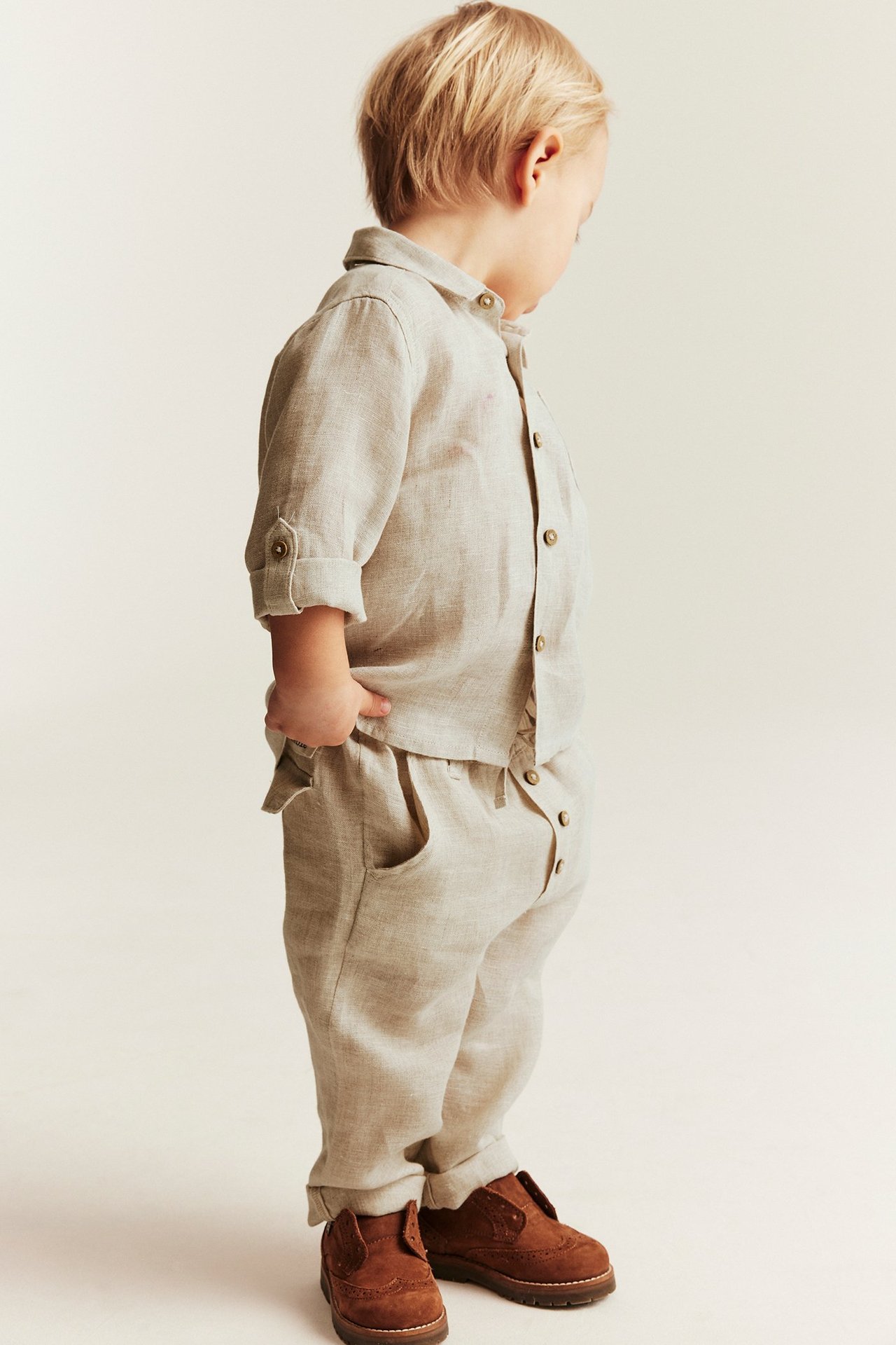 Side view of a kid wearing light beige linen drawstring trousers with rolled hems and a matching linen shirt.