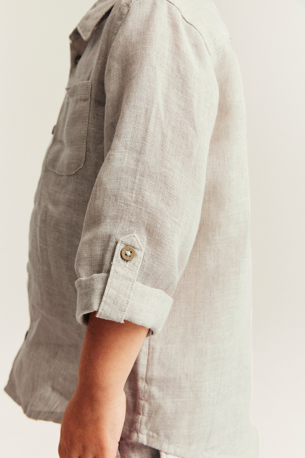 Close-up side view of a kid wearing a light beige linen shirt with rolled-up sleeves and a breast pocket.