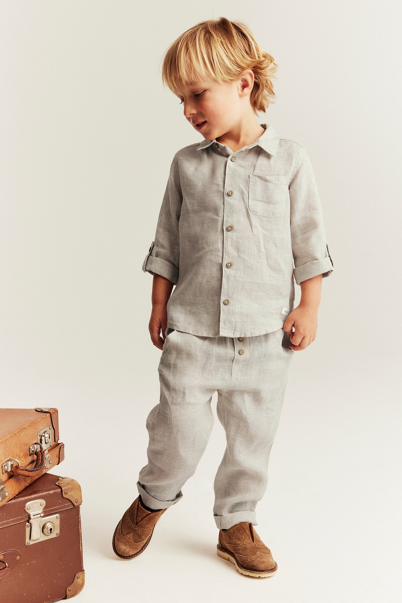 Front view of a kid in a light beige linen shirt with rolled-up sleeves, linen trousers, and brown suede shoes.