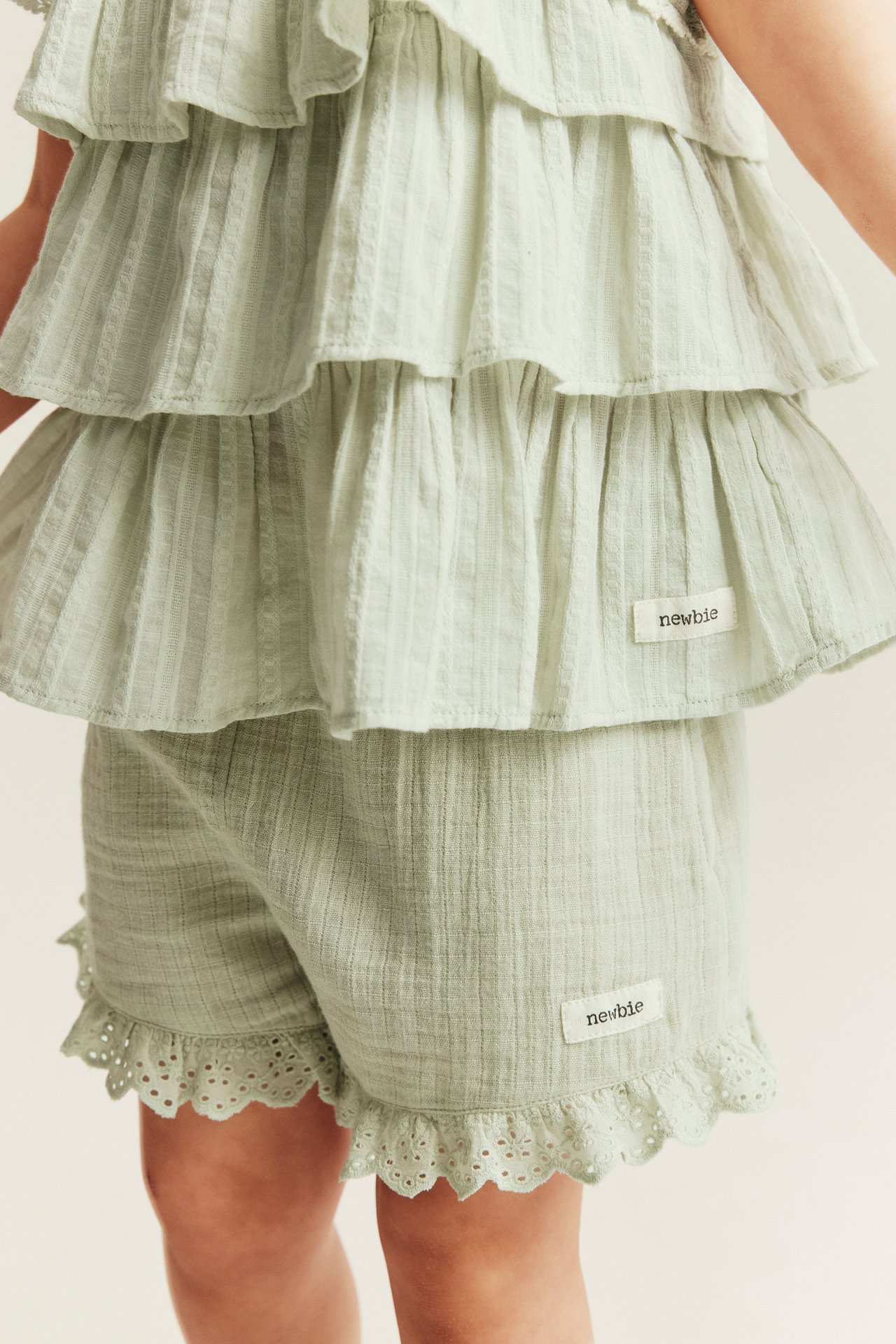 Front view of a kid wearing light green woven cotton shorts with lace ruffles, paired with a matching tiered ruffle top.