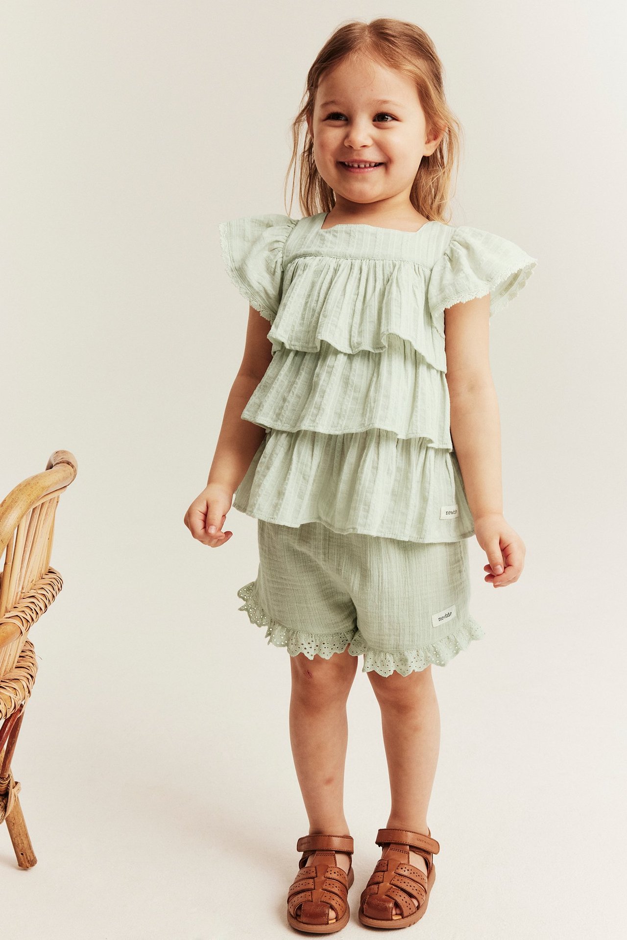 Front view: kid wears light green cotton shorts with lace ruffles, a matching tiered top, and brown sandals.